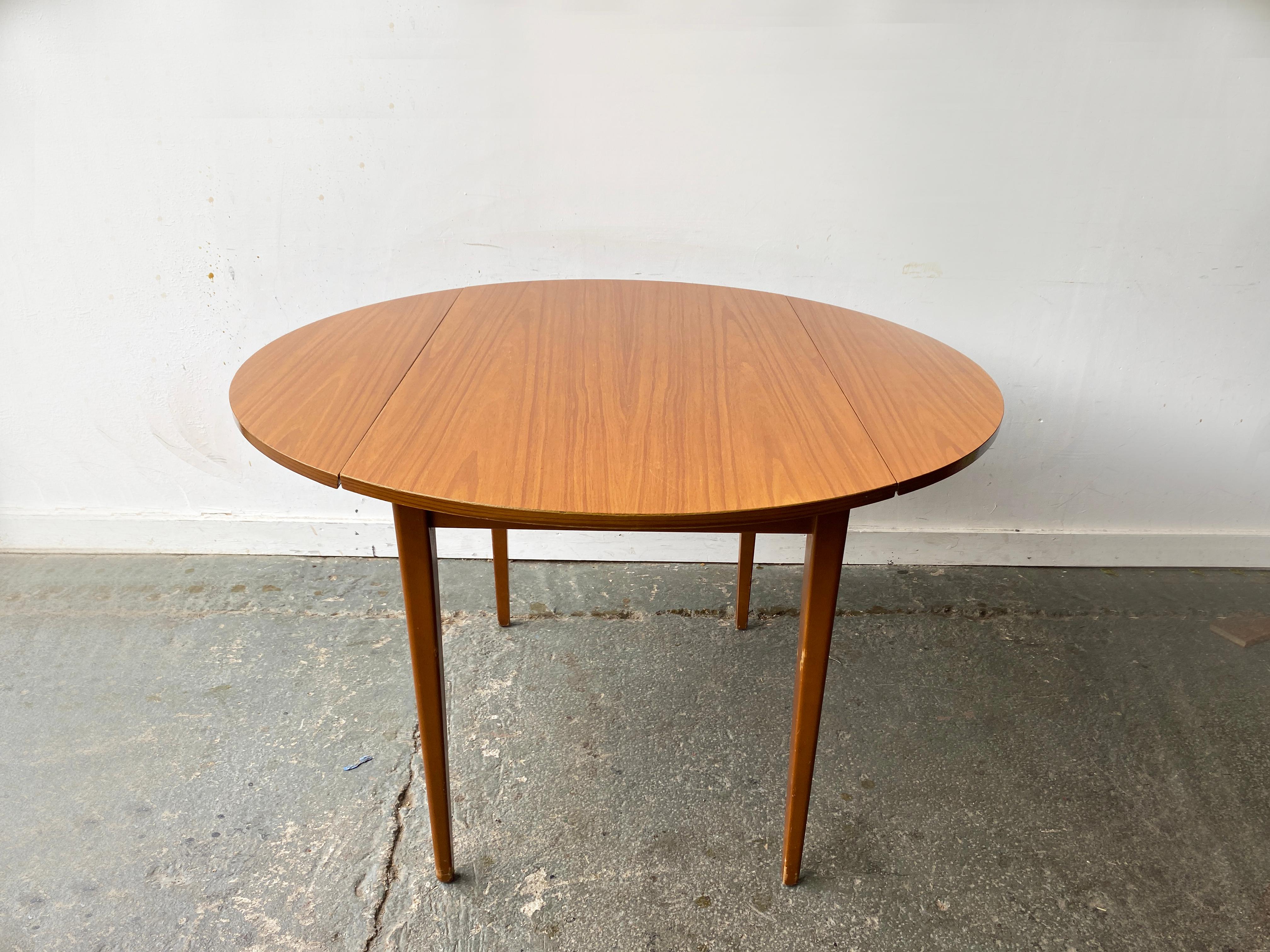A compact Mid-Century circular dining table with slim tapered solid Beech legs. The table top surface is finished in a wood grain effect Formica. The table features a clean, minimalist design with extendable leaves making it well suited for smaller