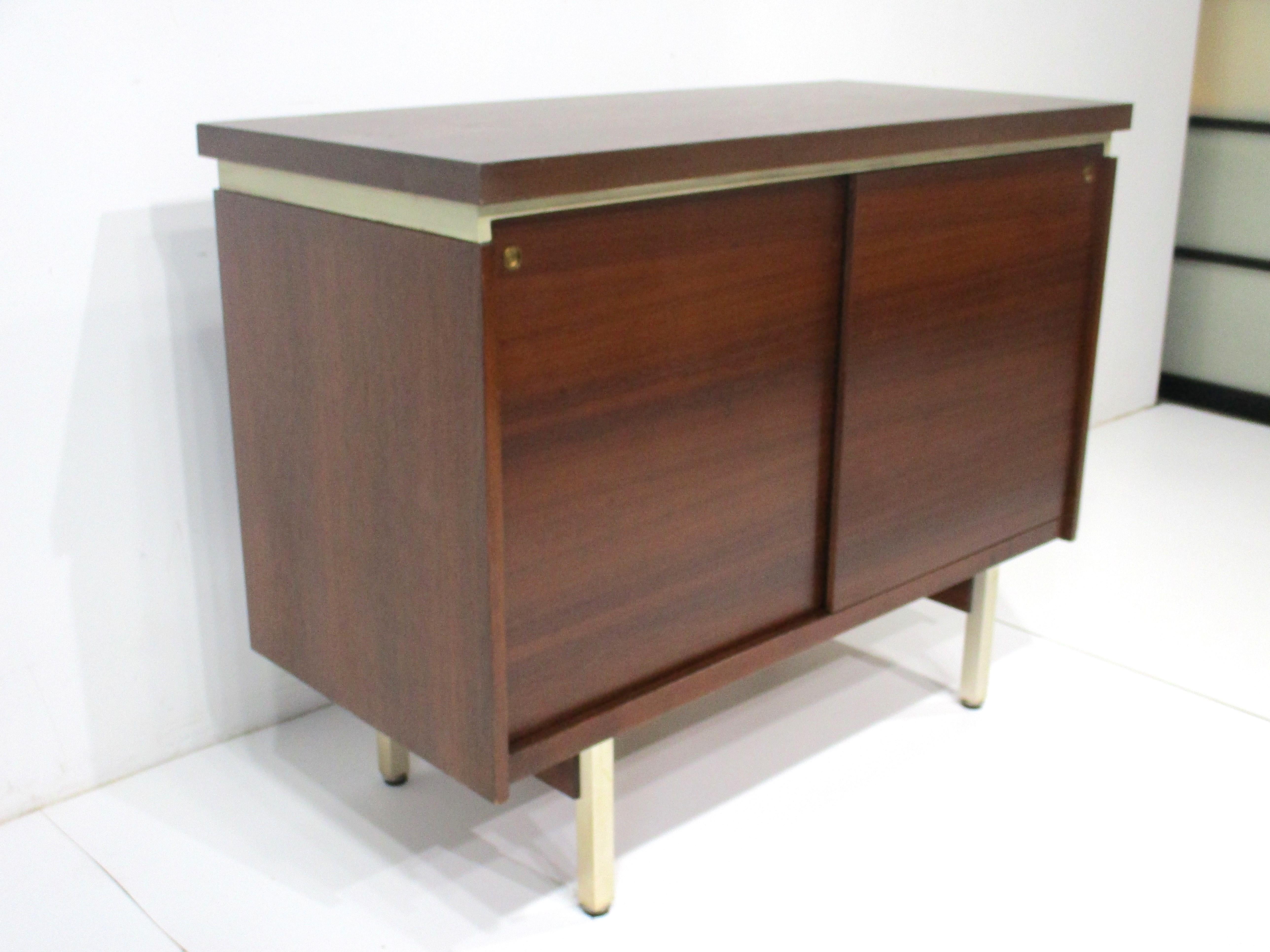 A small scale walnut two door credenza cabinet with two adjustable shelves sitting on gold brass toned metal legs . The piece has very nice graining of the wood front and on the finished backside . The top seems to float with a leatherette trim just