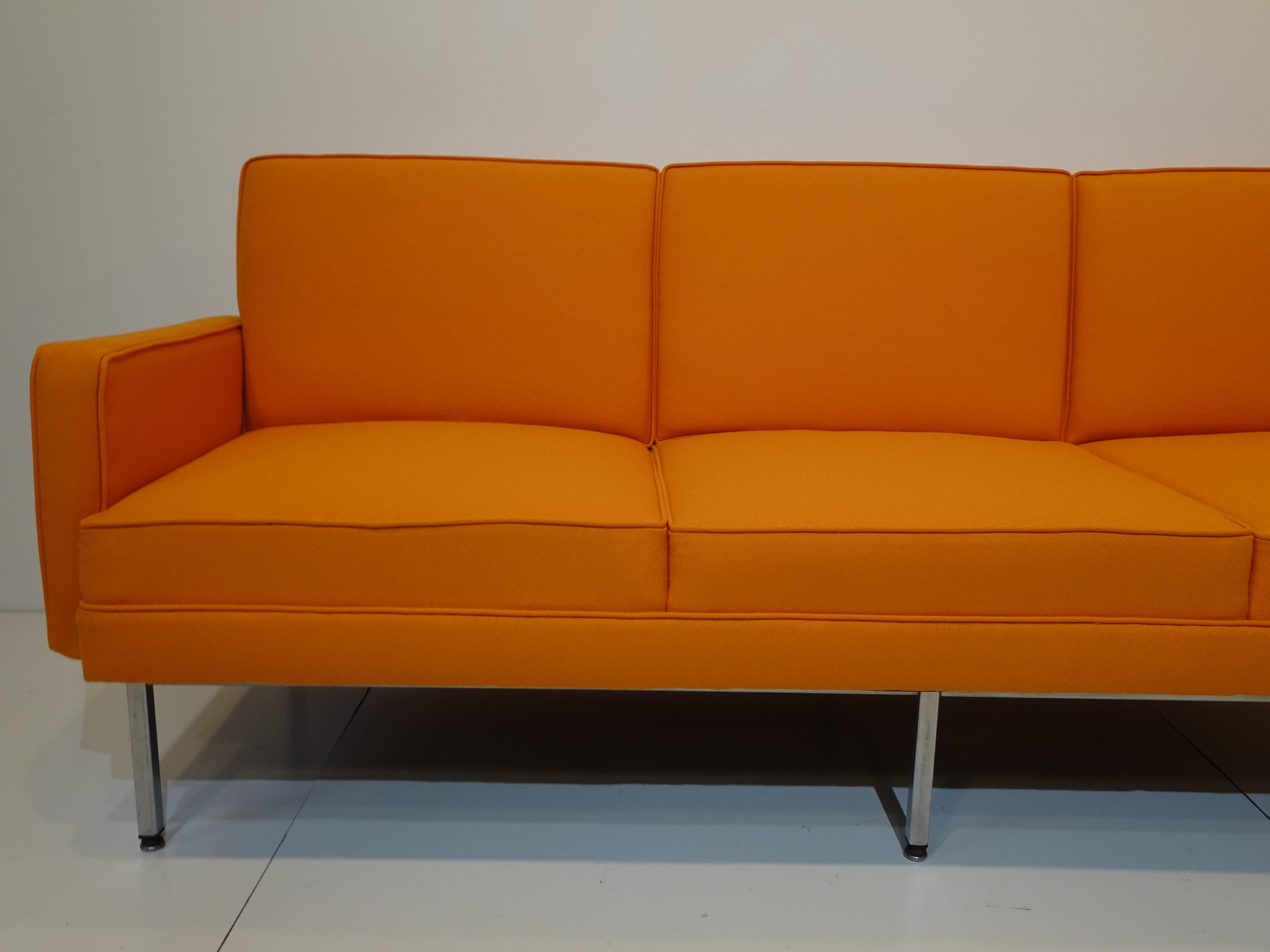 A steel framed sofa in a tight woven soft tangerine orange fabric upholstered with built in back and bottom cushions. Having arms to each side for comfort, sitting on aluminum steel legs with foot pads to protect your floors designed and