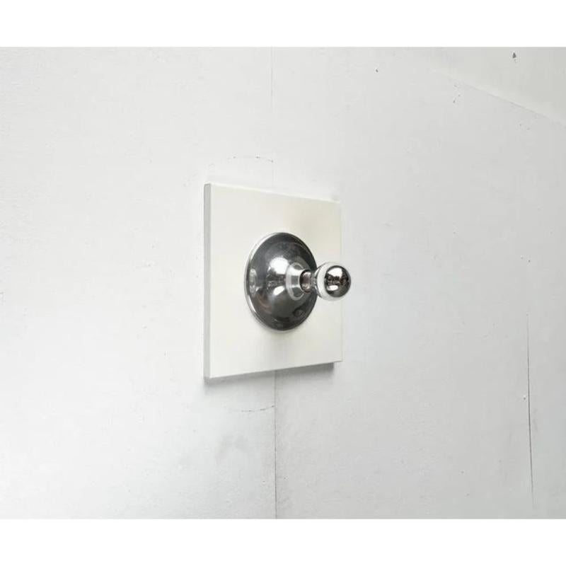 Imposante Raumzeitalter-Decken- oder Wandleuchte.

Die Lampe besteht aus zwei Elementen. Die quadratische Grundplatte und der angebrachte Rahmen. Die Grundplatte ist aus Metall gefertigt und weiß lackiert. Die Blende aus poliertem Aluminium verläuft