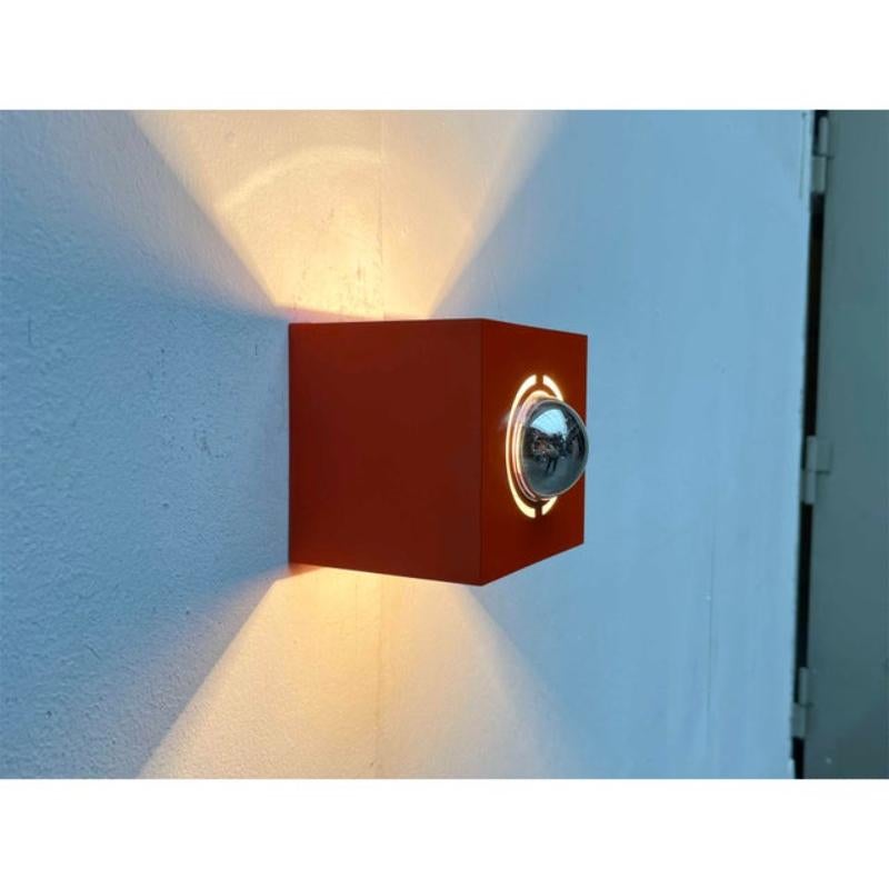 La lampada colpisce per la sua semplice forma cubica. La struttura metallica che funge da riflettore della lampada è rifinita esternamente in rosso-arancio. L'interno è bianco per garantire una migliore riflessione della luce. L'apparecchio è aperto