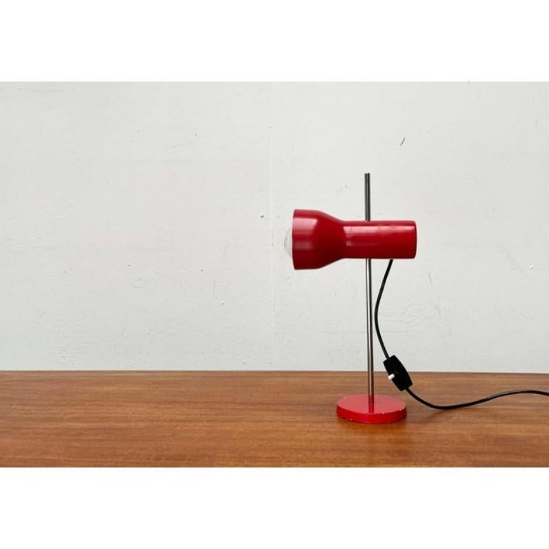 Un design minimalista. La testa della lampada è caratterizzata da una semplice forma a faretto ed è rifinita in rosso. Il colore del paralume si ripete nella base del piatto. La testa può scorrere su e giù per l'albero grazie a un semplice