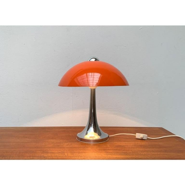 Lampe à poser en forme de champignon de l'ère spatiale.

La lampe présente une forme classique de champignon. Le grand abat-jour en métal est fini en orange et repose sur une base en forme de trompette. Il comporte une ouverture en haut au centre