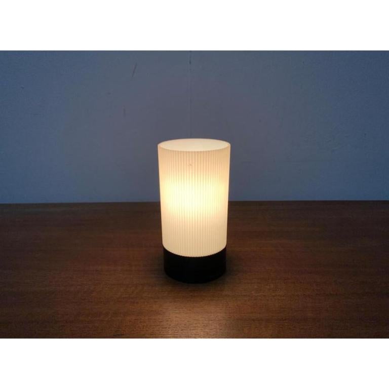 Mid Century table lamp by Erco.

A minimalist design. The lamp shows a simple cylindrical shape. It is divided into a base and the shade. The shade is made of translucent plastic and shows a grooved surface. The base is made of black plastic. A