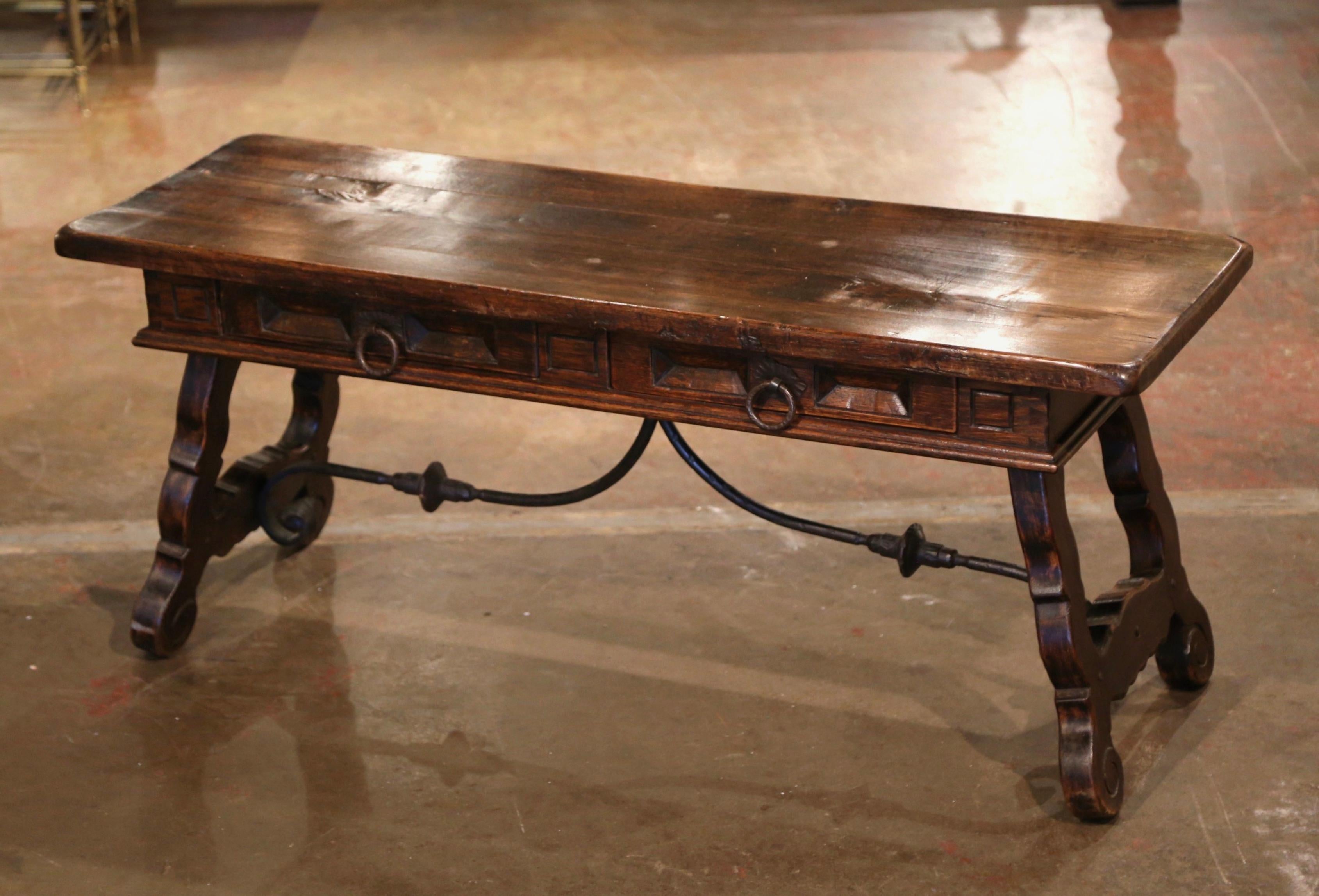 Crafted in Spain circa 1950, the rectangular antique fruit wood trestle coffee table stands on carved scrolled legs joined together with a forged iron stretcher over two drawers dressed with iron pulls; the top features rounded edges and corners.