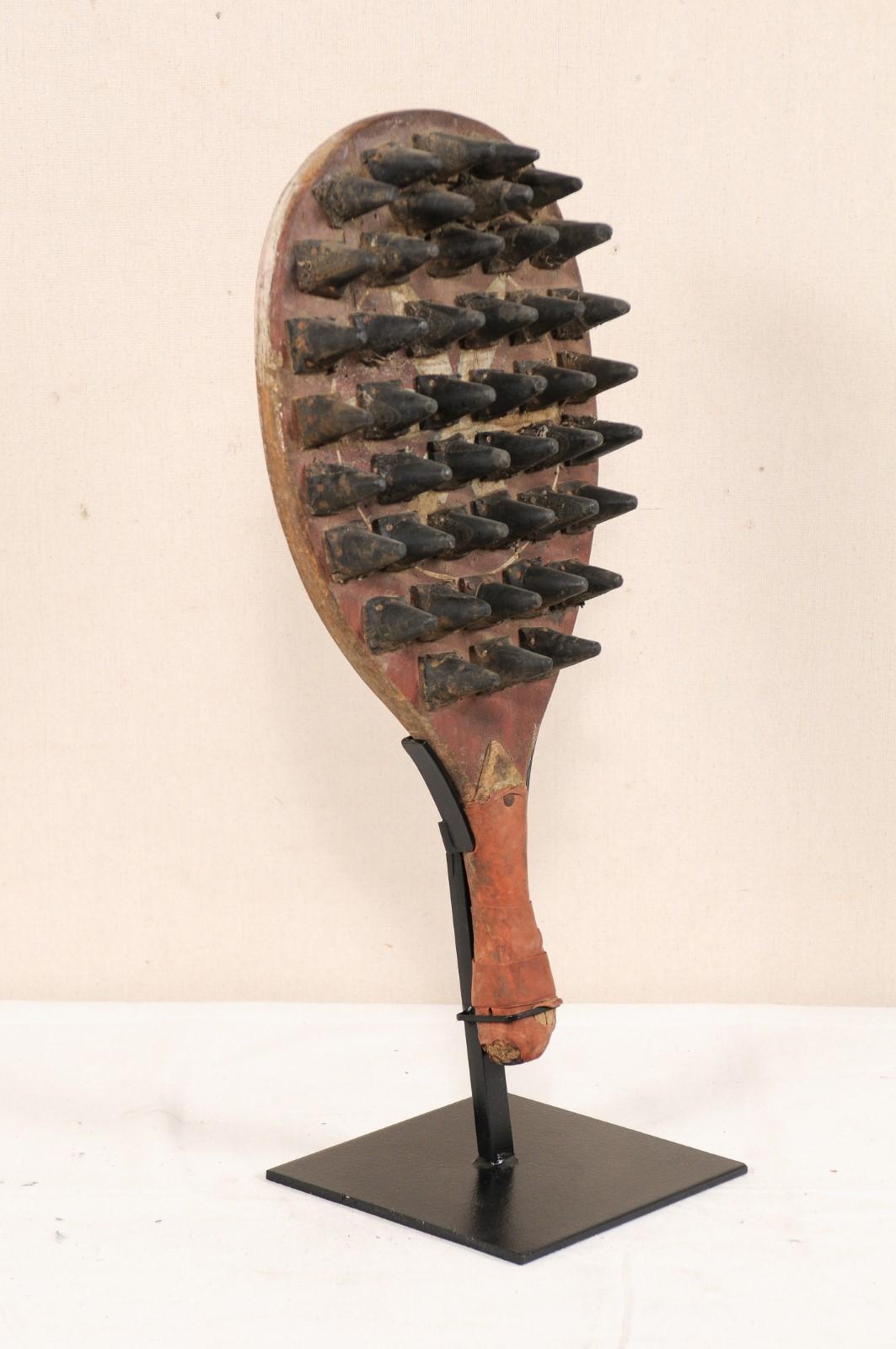 A wooden spiked gaming paddle from the mid-20th century. This vintage game paddle from India features a wooden handle and oval-shaped body with spikes projecting out at one side, with remnants of its original decorative paint showing throughout