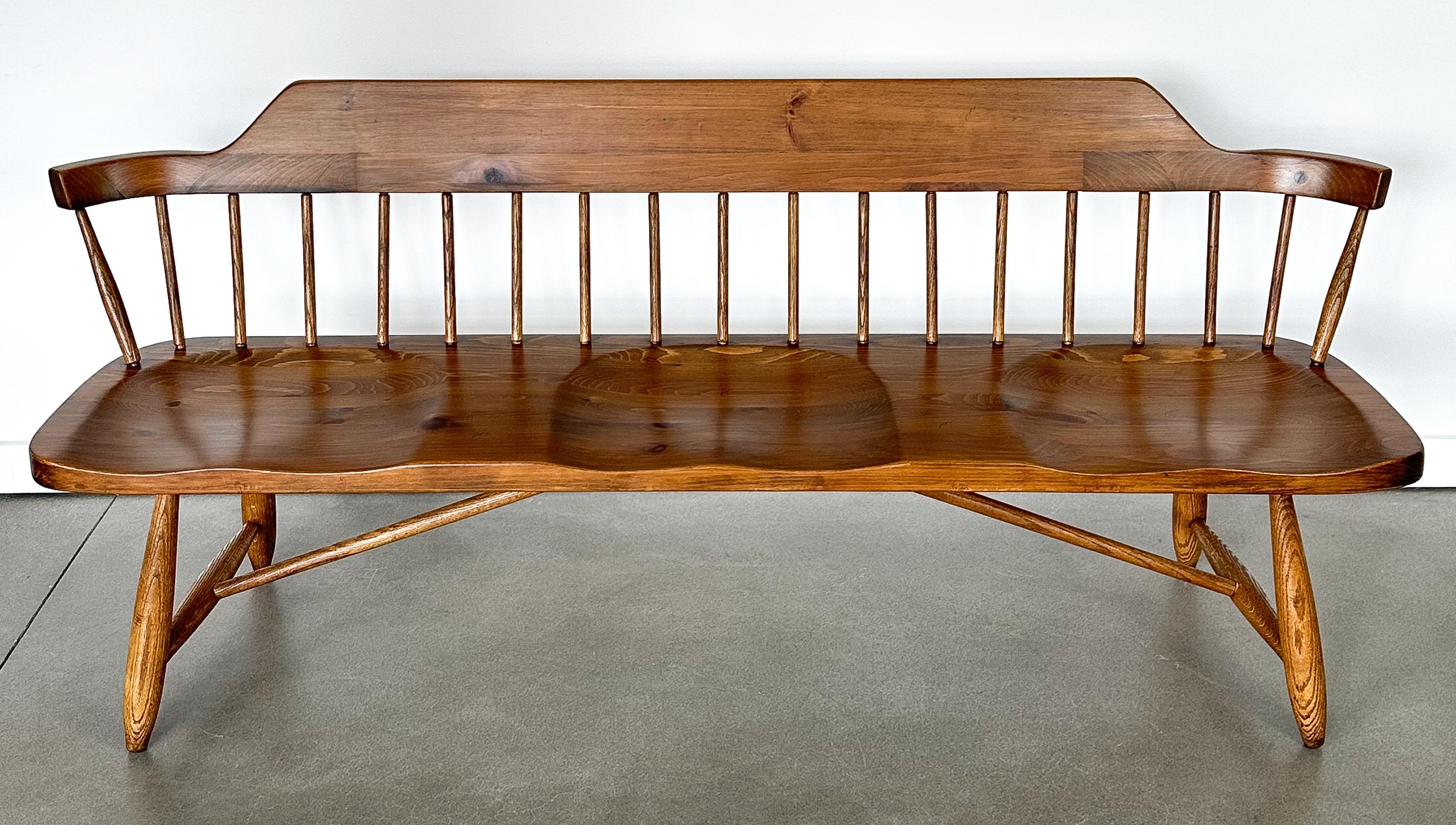 A mid-century spindle back three seat bench by Conant Ball, circa 1950s. The seat and back are constructed from solid pine and are stained in a medium walnut tone. Legs are solid oak. The seat features three individual sculpted seats. Backrest has