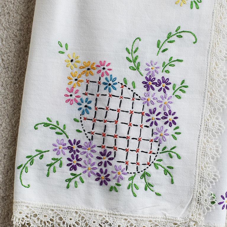 A pretty and colorful midcentury hand-embroidered tablecloth. The piece is square and created from linen. Crotcheted lace decorates the edges in a flower pattern. The corners of the table covering feature pineapples in a rainbow of colors.