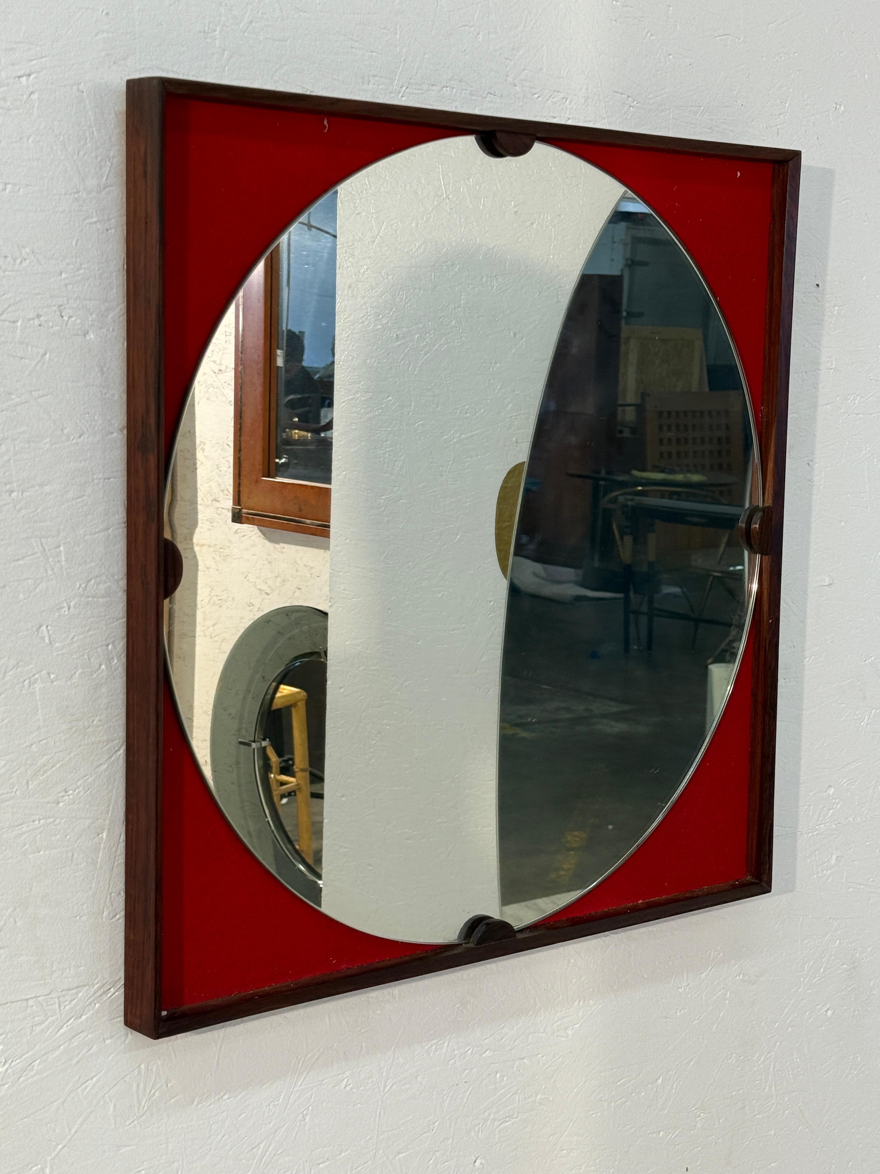 Italian mid-century modern wall mirror features a bold square wooden frame with a vibrant red background. The circular mirror insert is accented by four wooden brackets at the edges, creating a geometric interplay between square and circle. Its