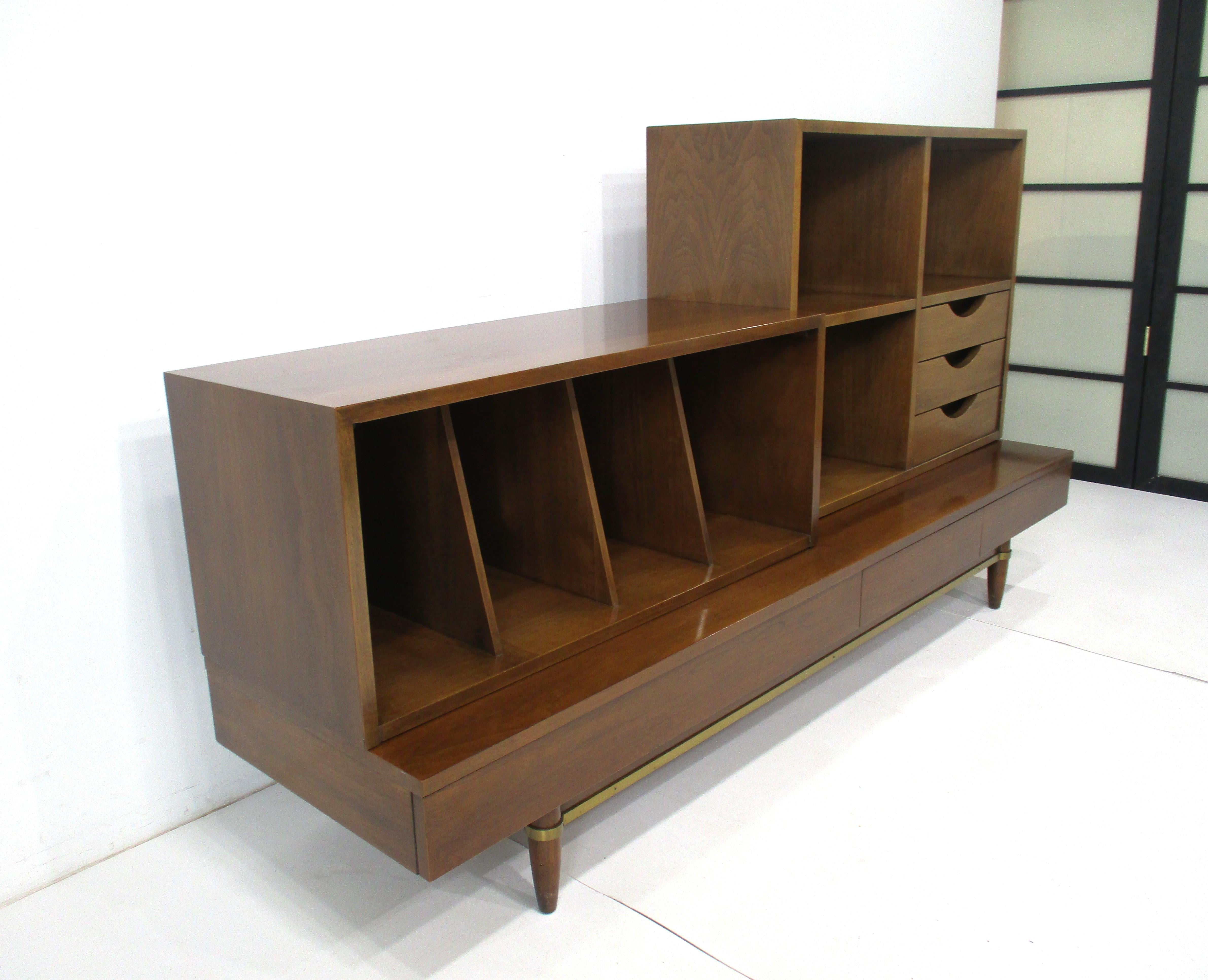 A three piece walnut Mid Century stereo record cabinet on a platform styled base with three drawers . The left upper cabinet has four record bins and the right side cabinet has three storage or bookcase bins and three drawers , each can be moved on