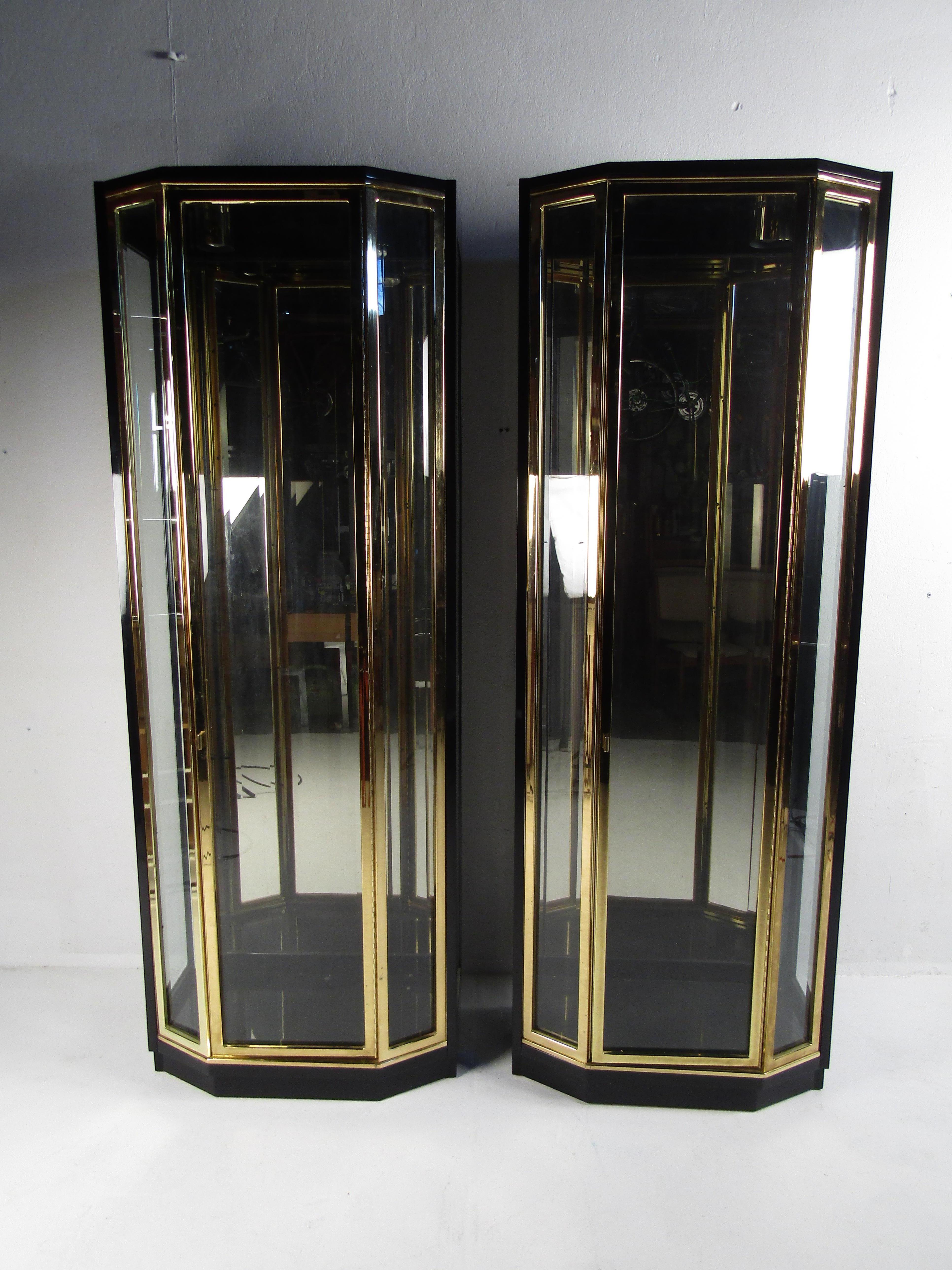 This elegant pair of vintage modern curio cabinets feature two cylindrical lights on the top within a uniquely shaped design. Elegant with brass trim, black lacquered casing, and glass fronts adding to the allure. A tall cabinet door opens to unveil