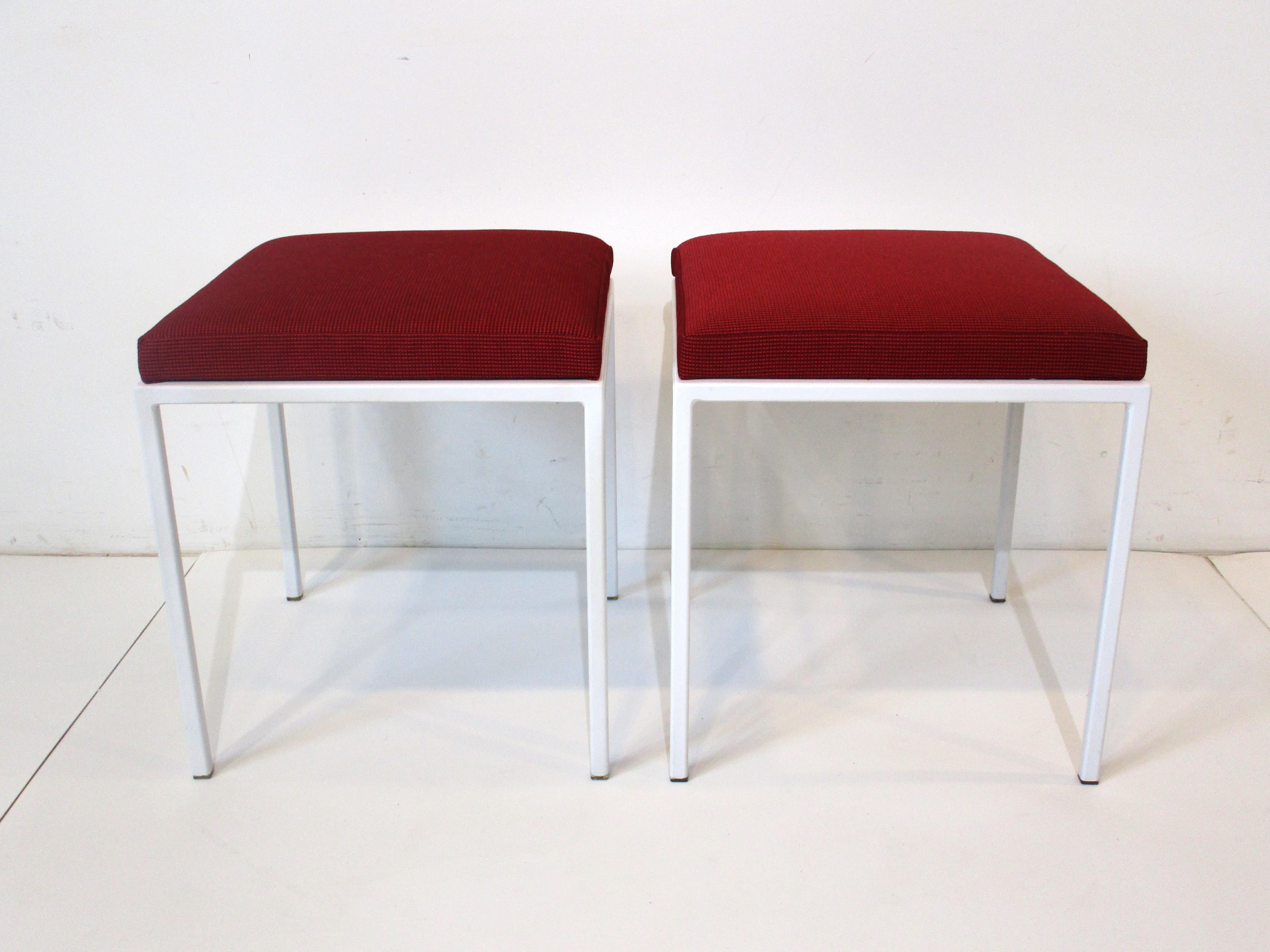 A pair of smaller sized midcentury steel framed stools in satin white with clear plastic foot pads to protect your floors . Upholstered in a dark red pattered farbic from Knoll with nice cushioning designed by D.R. Bates and Jackson Gregory Jr for