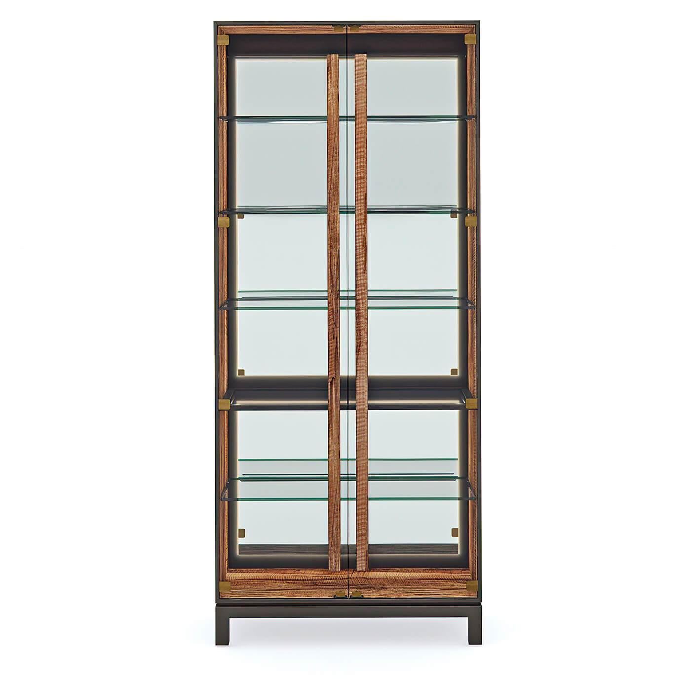 A Mid-Century-style glass door display cabinet illuminates curated displays and collectibles. Finished in Greenway, a complex mix of dark brown, black, and olive tones, the case exterior offers rich contrast to the door handles and interior, with a