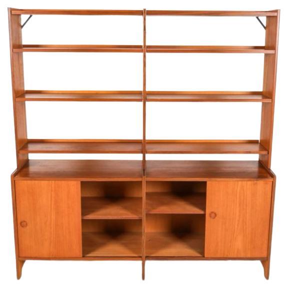 Mid century Swedish teak bookcase wall unit with lower credenza cabinet. Double unit with lower credenza with sliding doors that has open backed shelves on top. Lower credenza has round inset handle with adjustable shelves inside. Shelving is