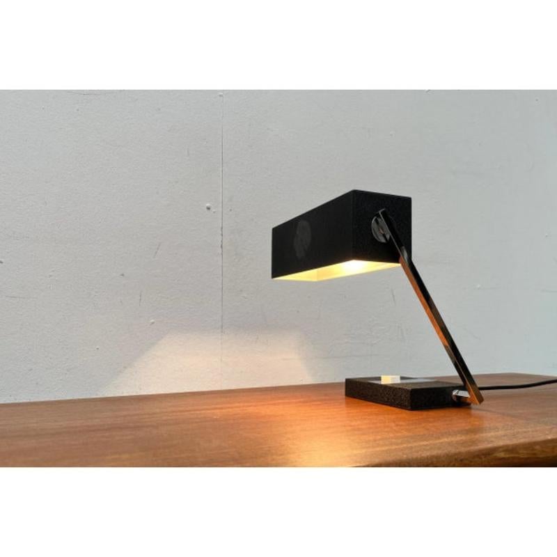 Minimalist mid-century table lamp from the Swiss manufacturer Megal.

Cubic shapes run through this design, from the shade to the arm to the base to the switch. The rectangular lamp head sits at the end of an angled arm. It is movably mounted via