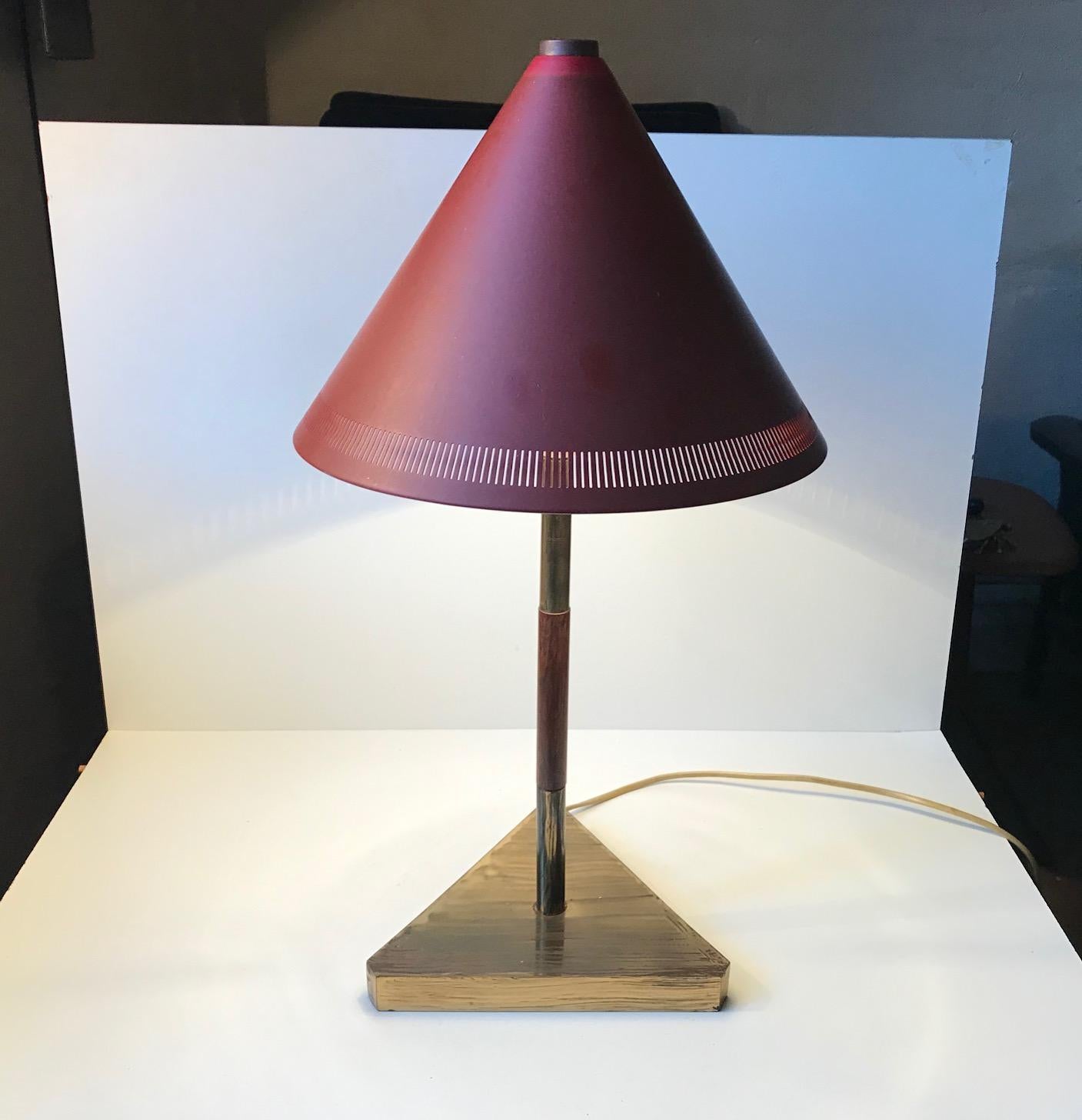 This Danish table lamp has a conical-shaped shade and a triangular brass base. It was manufactured by Lyfa in Denmark during the 1960s. The piece features a brass base and stem and a centerpiece in teak. The perforated clay-red shade is made from