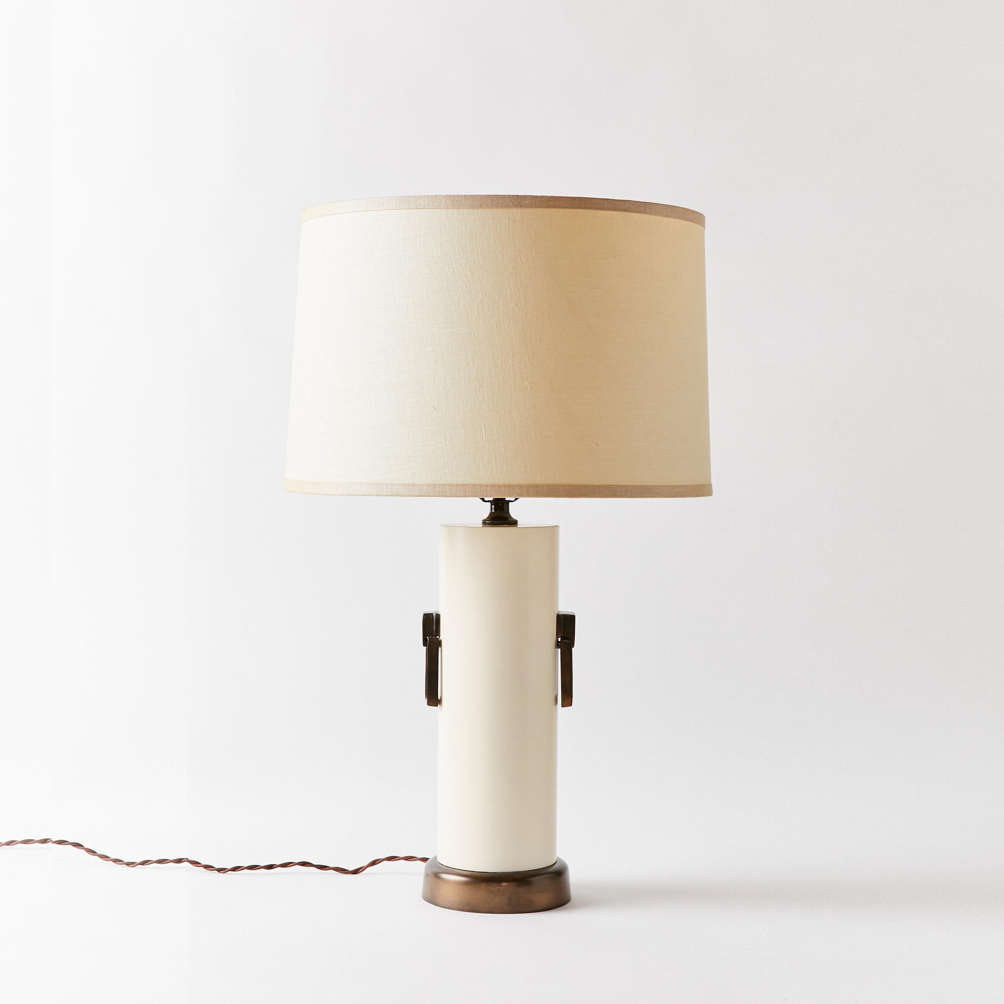 Cylindrical shape table lamp finished in off white and siting on an antique bronze base. Rectangular pull rings in antique bronze show on each side of the body.