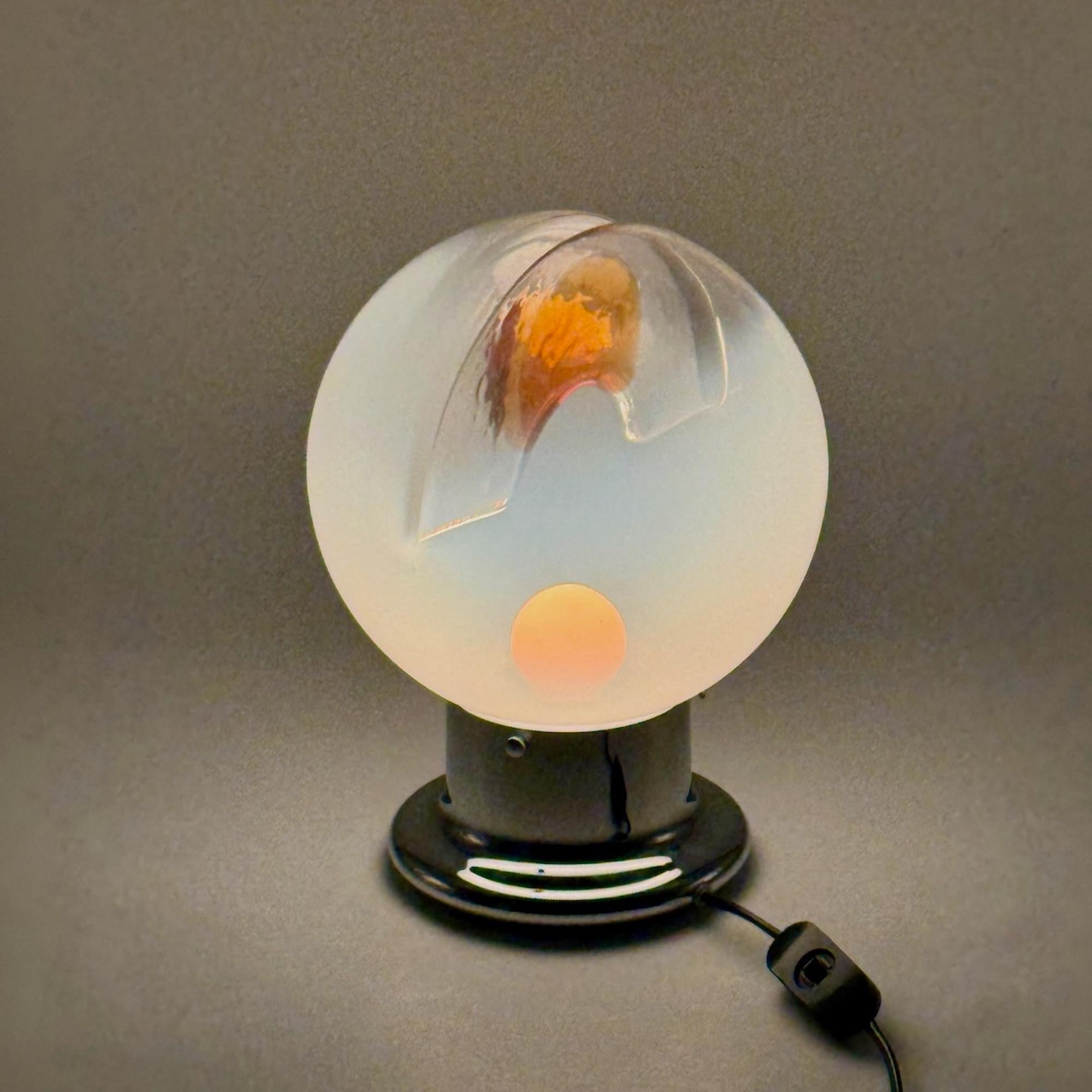 Esta lámpara de los años 70 es un notable ejemplo de diseño de iluminación vintage, con una pantalla de cristal con un marcado efecto de división y una inclusión naranja en el interior, que crea un sorprendente juego de luz y color.

La base