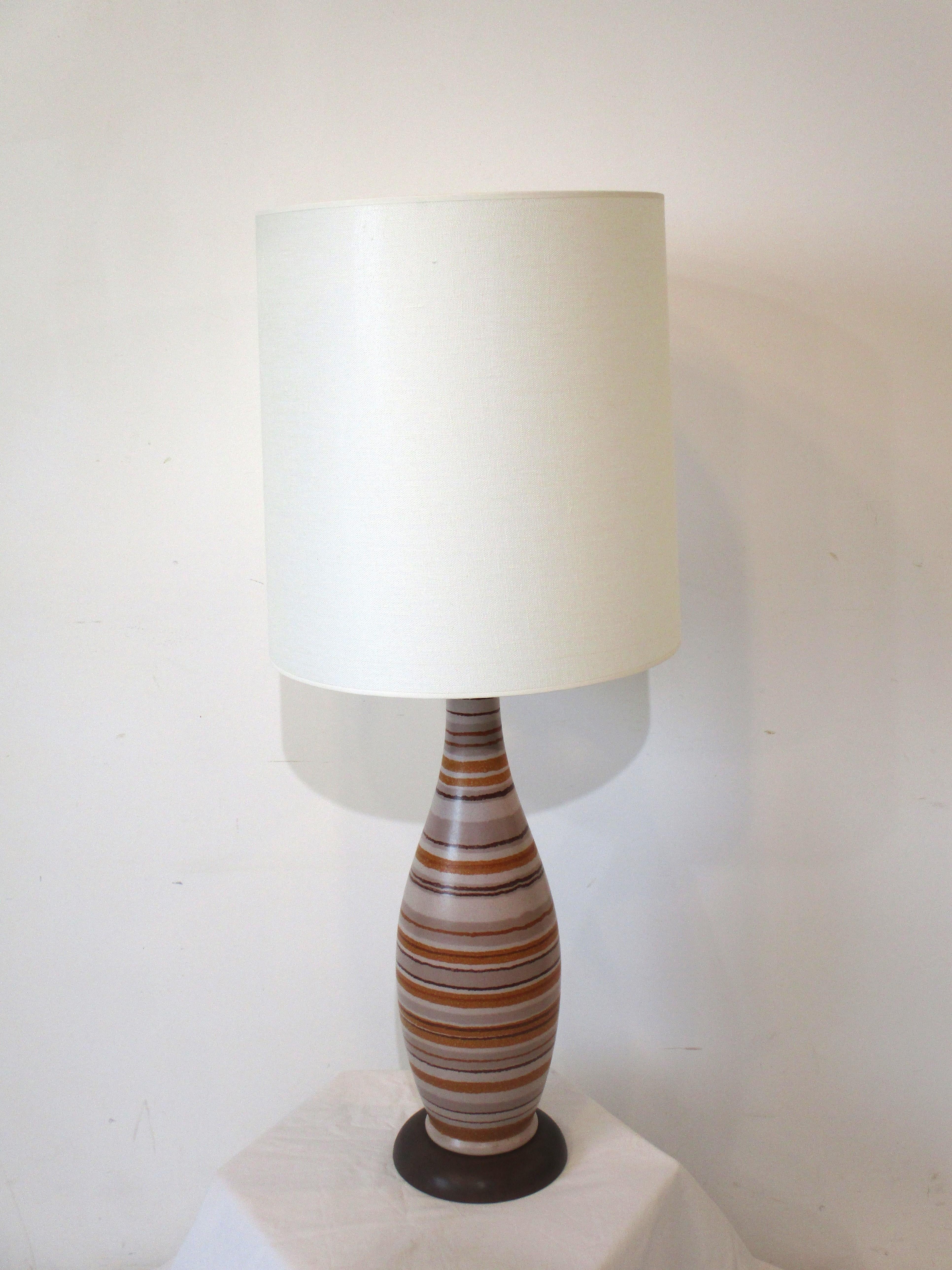 A midcentury ceramic table lamp with beige colored body having lines of burnt orange, browns and tans. Sitting on a dark walnut toned wood base topped with a cream colored linen shade manufactured in the manner of Bitossi Italy. A visually well