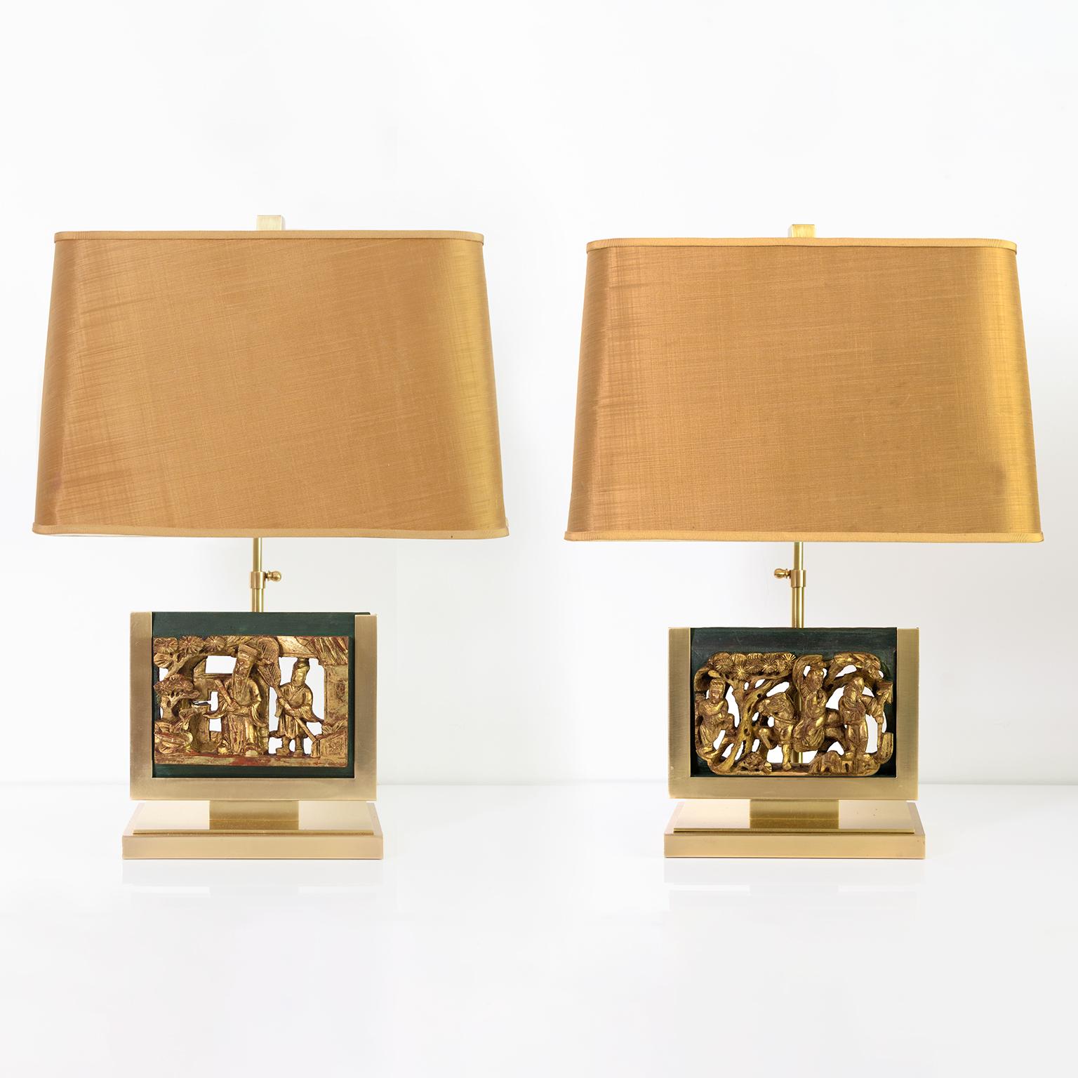 Pair of mid-century table lamps in polished solid brass with mounted Asian gilt-wood carvings. Each carving depicts a scene with figures in a landscape. The original fabric and paper shades show wear but have perfect proportions and sit on an