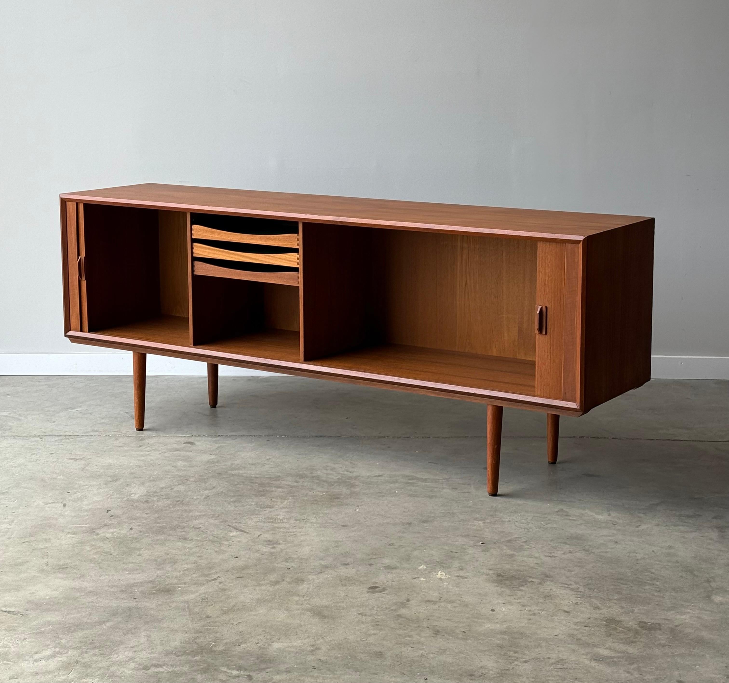 Credenza di metà secolo progettata da Svend Aage Larsen, Danimarca. Questa credenza monumentale ha un profilo elegante con due ante scorrevoli a tamburo con maniglie scolpite e poggia su alte gambe affusolate.

Composto da Teak abbinato a libro con