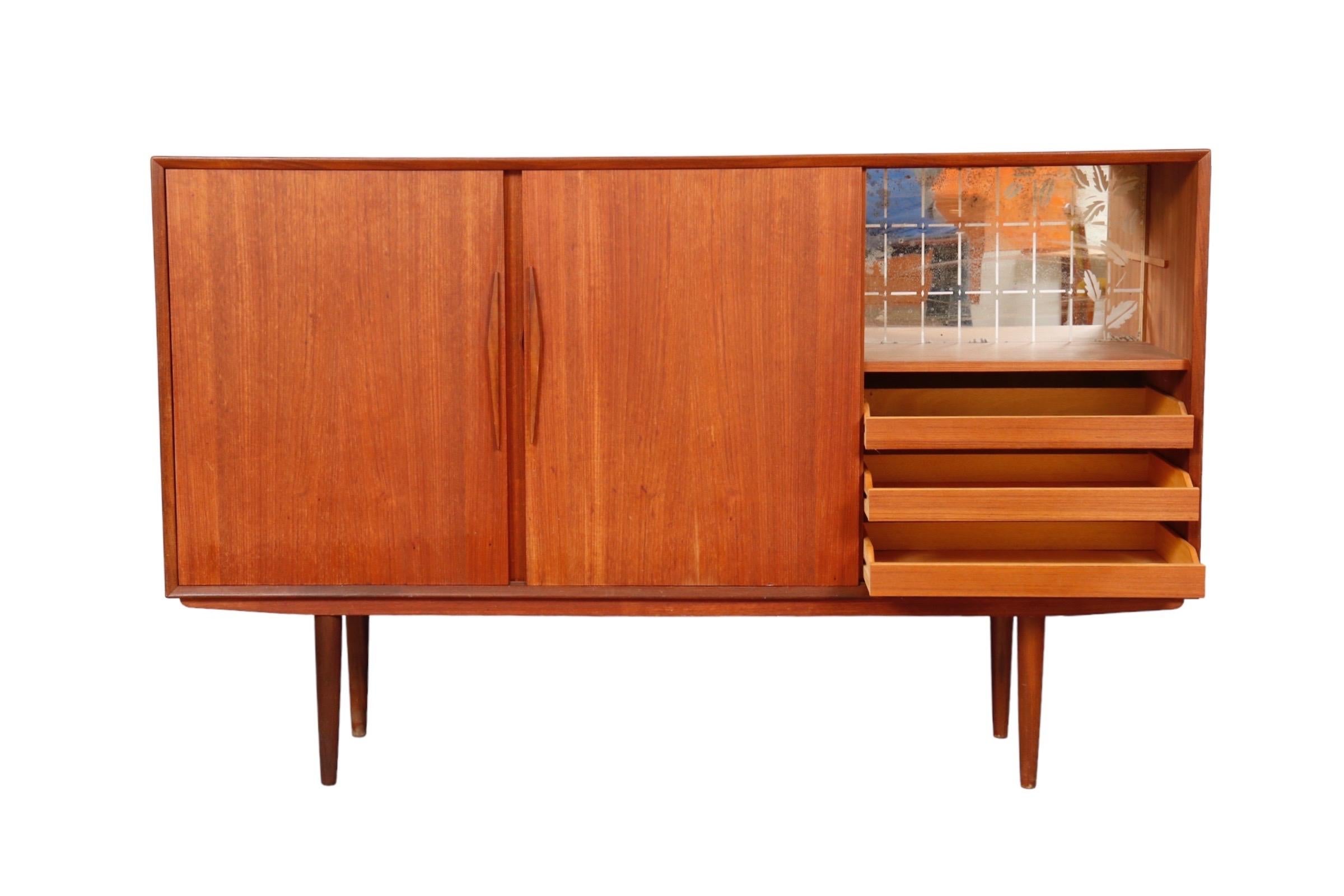 A Danish mid century bar buffet in teak. Three sliding doors reveal storage shelves left and center, and a mini bar area and three drawers to the right.