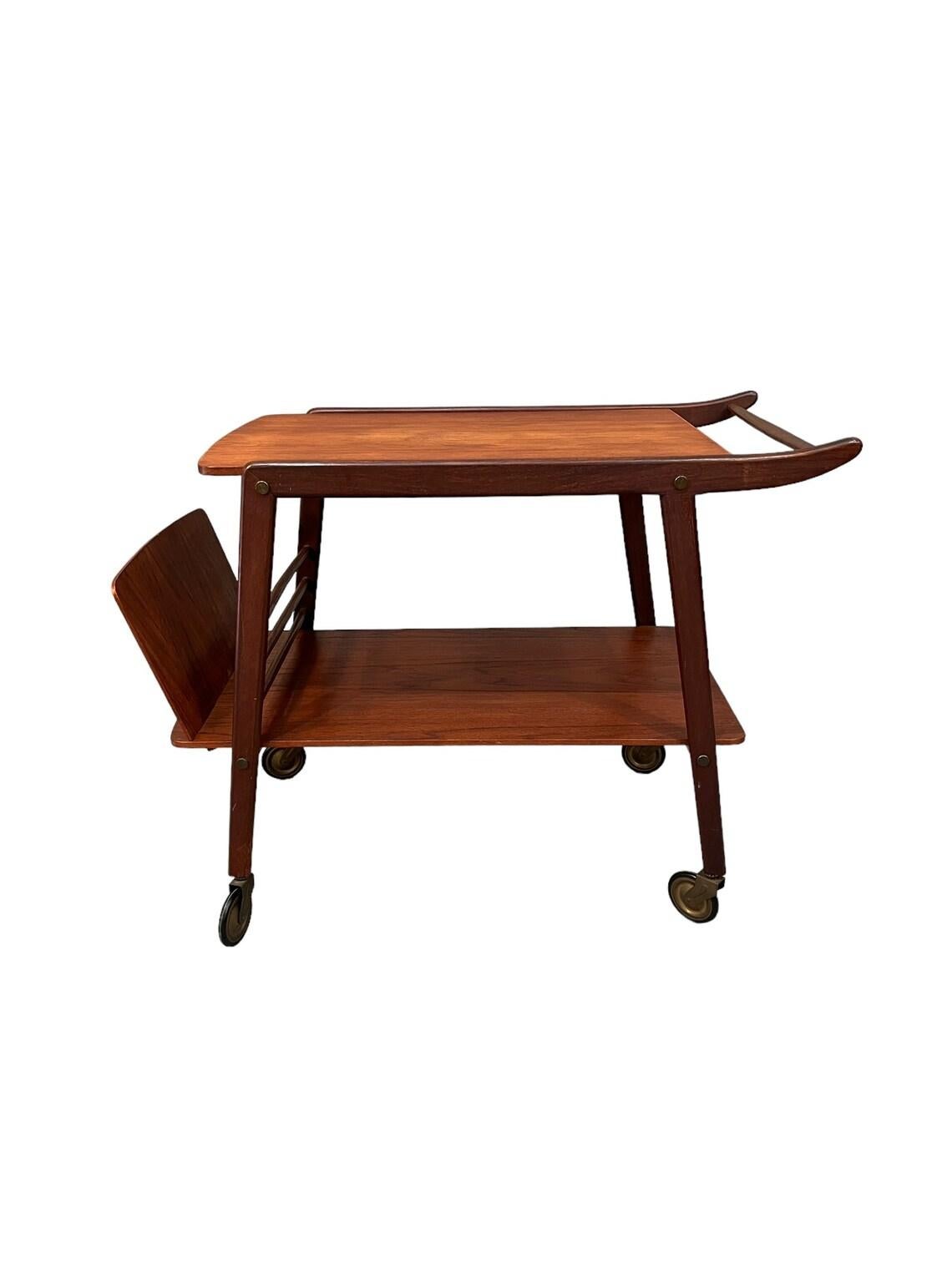 This Mid-Century Danish teak bar cart with magazine holder, circa 1960, is a beautiful example of Scandinavian design. Crafted by Hans Wegner, it features a medium wood tone, measuring 26.25 inches tall, and 20.5 inches wide, with a depth of 29.5