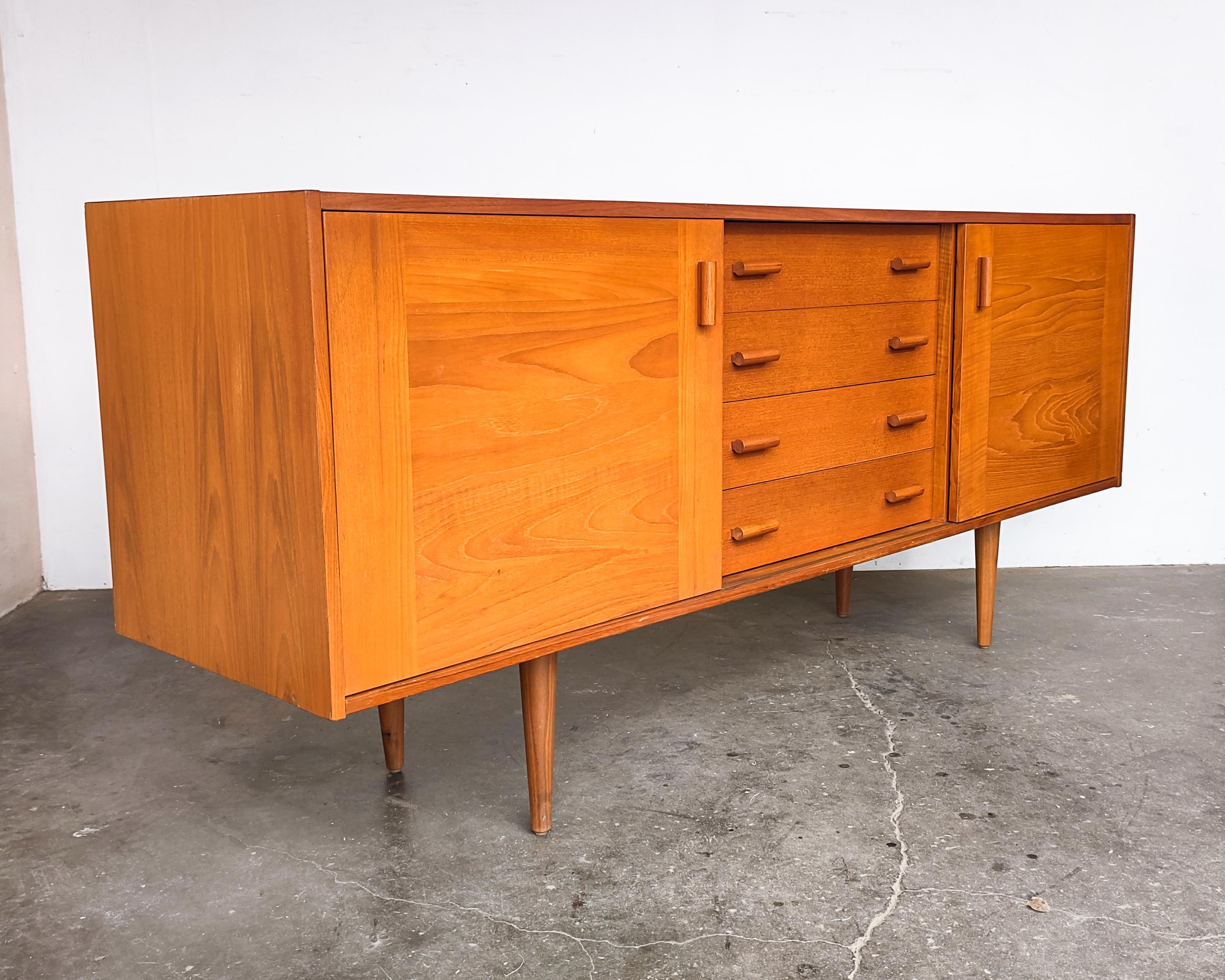Danish teak credenza by Domino Mobler with four drawers down the middle and sliding doors covering cabinets on either side. Each cabinet has an adjustable shelf and there is a hole for cables in the right-side cabinet. Teak dowel handles and drawer