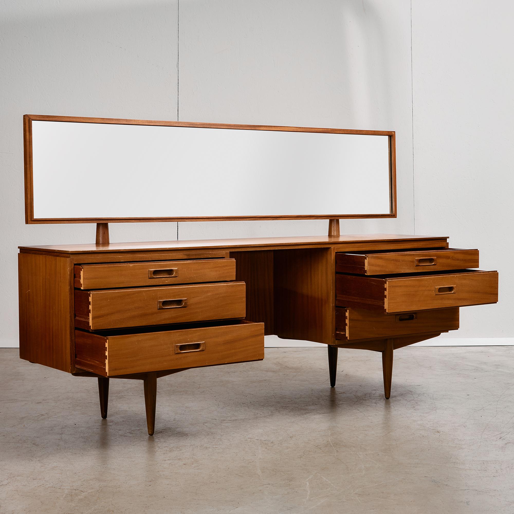 Este tocador de madera de teca de los años 60 es todo elegancia tranquila y detalles ingeniosos. La silueta es limpia y escultural, con un tono cálido y sutiles curvas que resaltan la belleza de la madera. Su característica más destacada es el