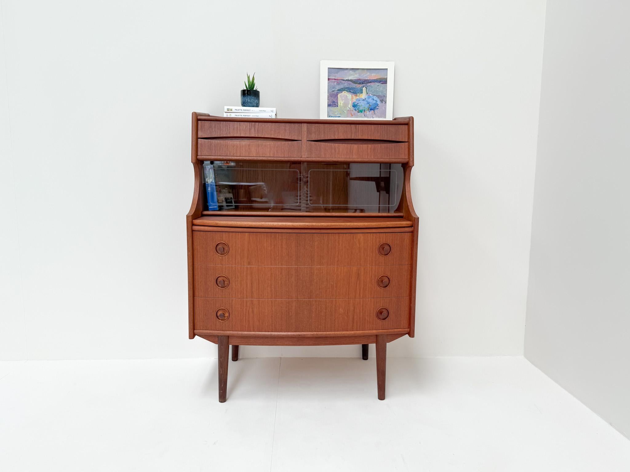 Este elegante escritorio de secretaria vintage está hecho de cálida madera de teca y presenta un diseño moderno atemporal de mediados de siglo. Ofrece una combinación práctica y elegante de almacenamiento y espacio de trabajo. En la parte superior,
