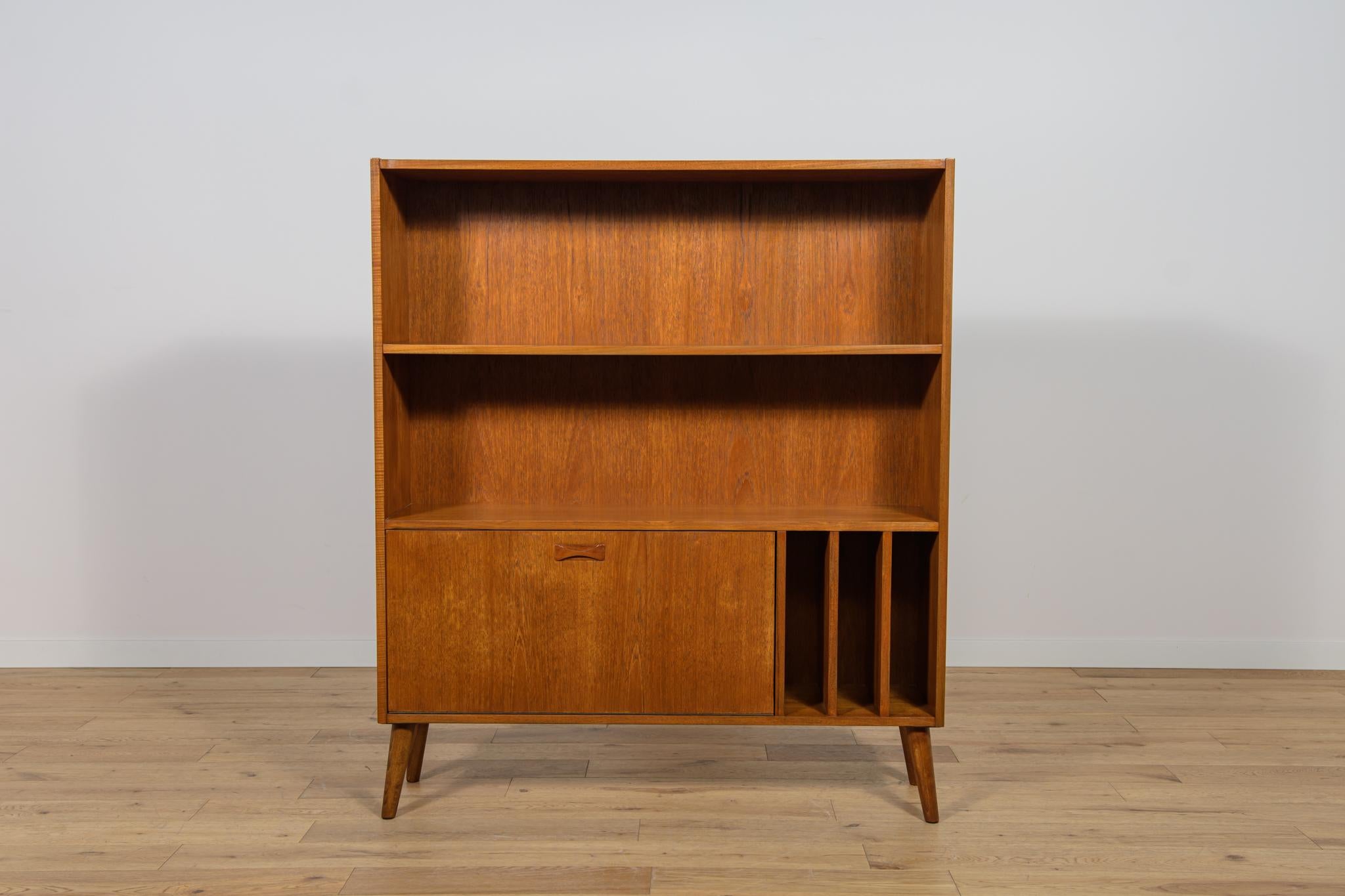 This bookcase made of teak wood in Denmark in the 70s. In the lower part there is a bar and a shelf for gramophone records, with profiled handle, there are shelves in the upper part.
Bookcase placed on slanting legs. The bookcase after a
