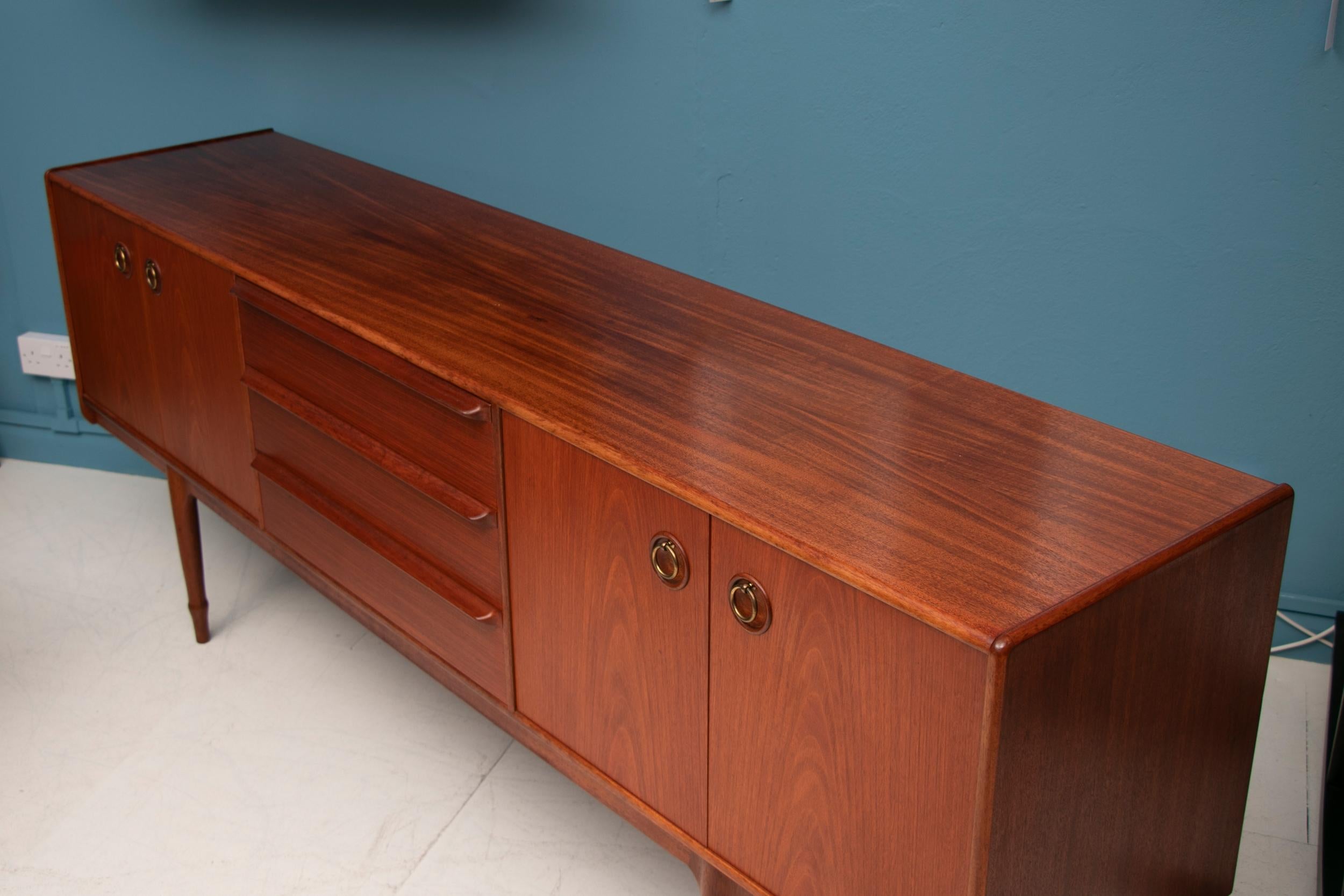 A stylish and functional midcentury teak sideboard with three central drawers flanked with two large cupboards, inset brass ring handles and beautiful carved tapered legs by McIntosh, circa 1970s.