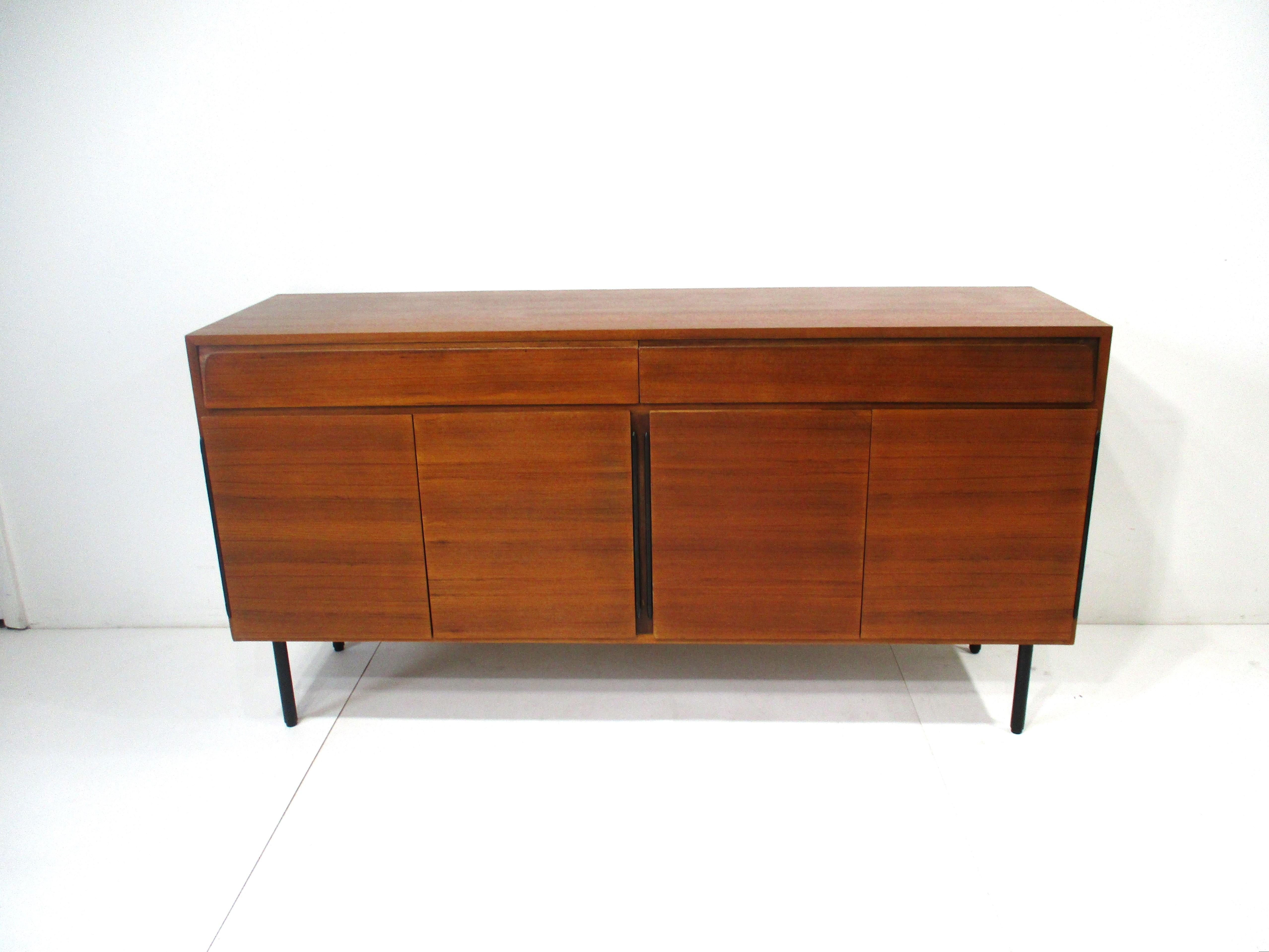 A teak wood credenza / sideboard with four doors , storage and adjustable shelves in a lighter wood . Having two drawers with one open and the other with dividers for separate compartments and detailed with satin black hinges and black metal legs .