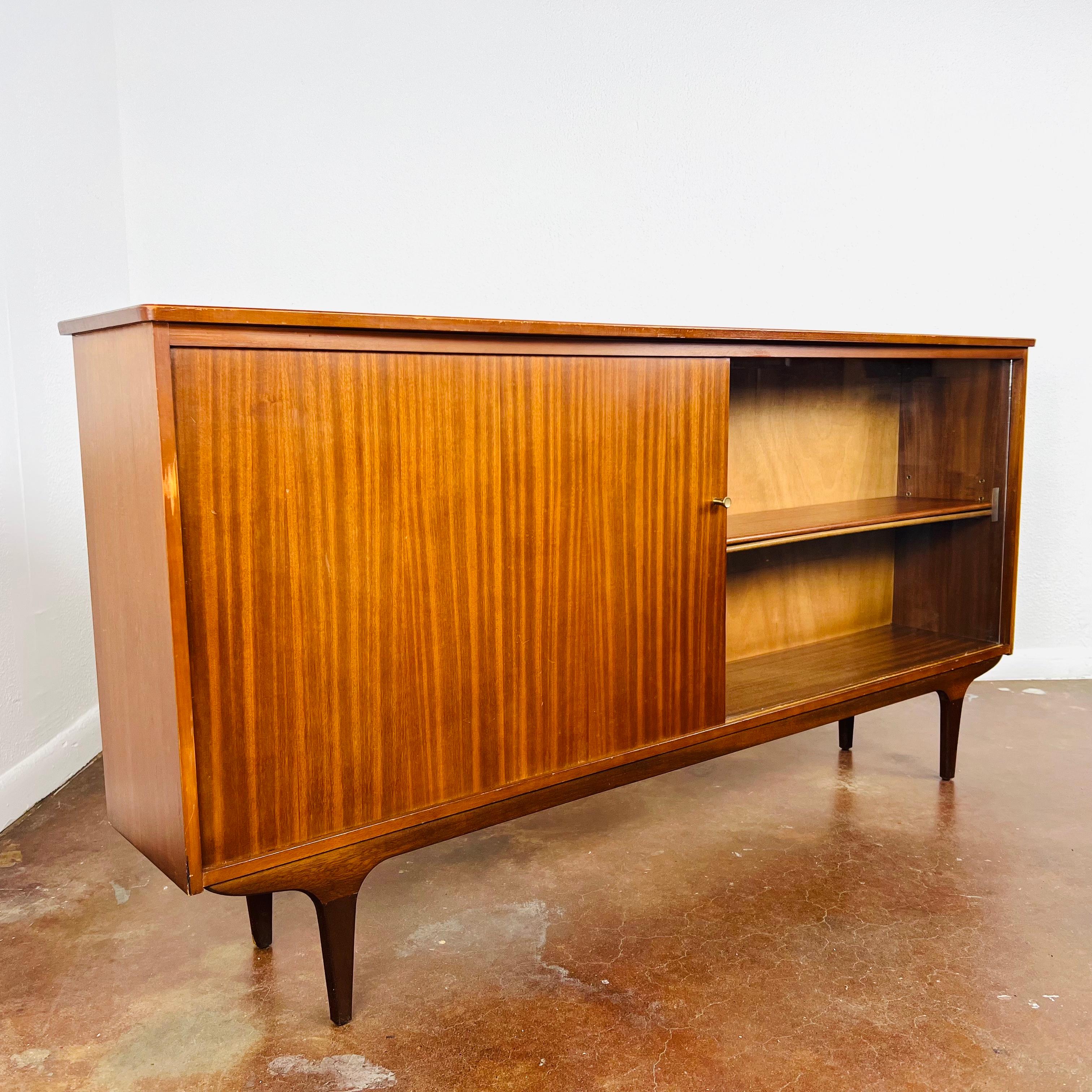 Mid-Century teak glazed sideboard with single-glass sliding door and single-wooden sliding door. Shelved interior, pin legs. Good, sturdy condition with minor surface marks/scratches due to age and use.

Dimensions:
30.25