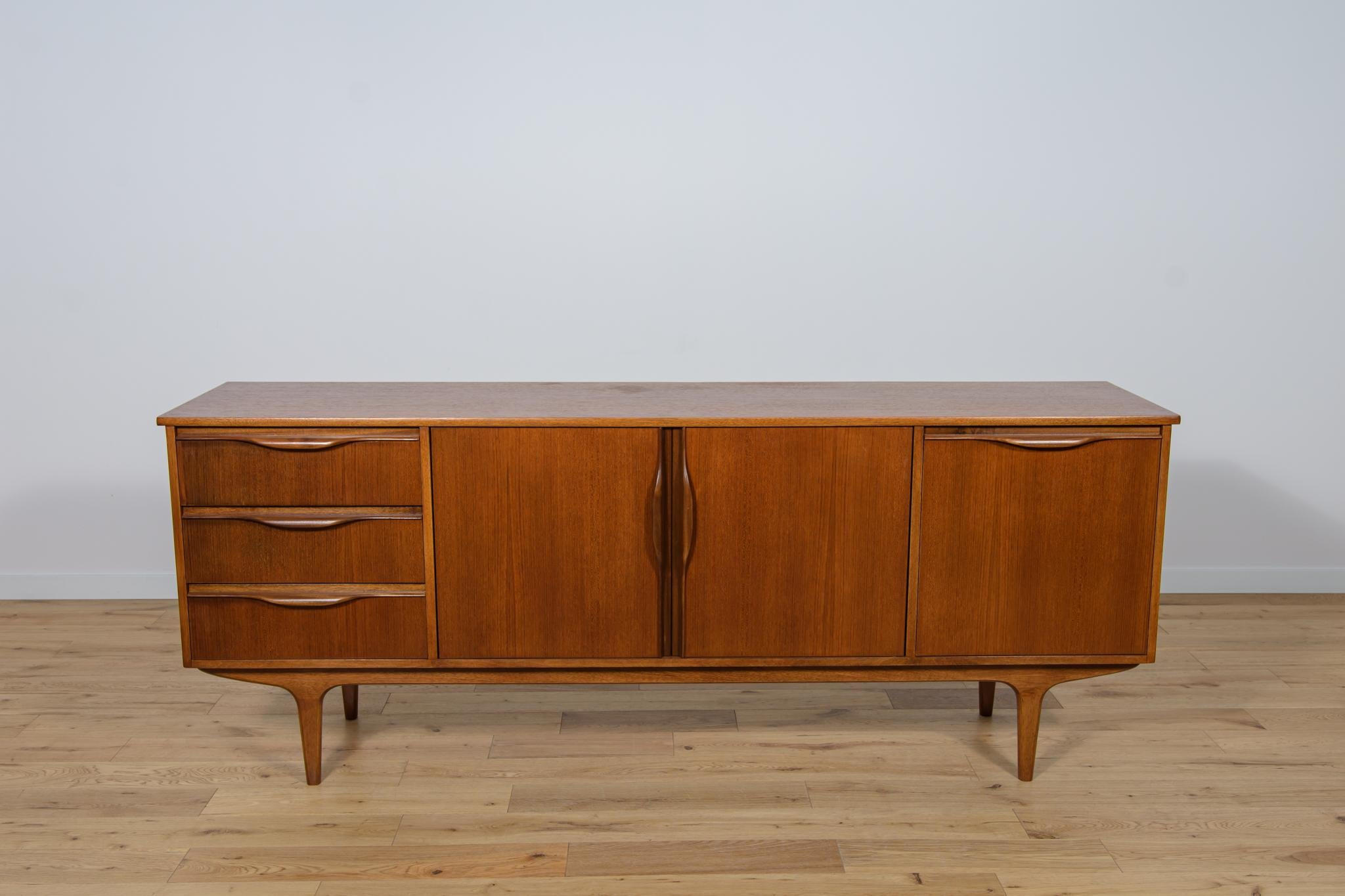 Sideboard produced in the 1960s in the British Jentique manufacture. The sideboard consists of three drawers and a cabinet with two doors and a bar. The sideboard has contoured solid wood handles. Sideboard made of teak wood. The sideboard has been