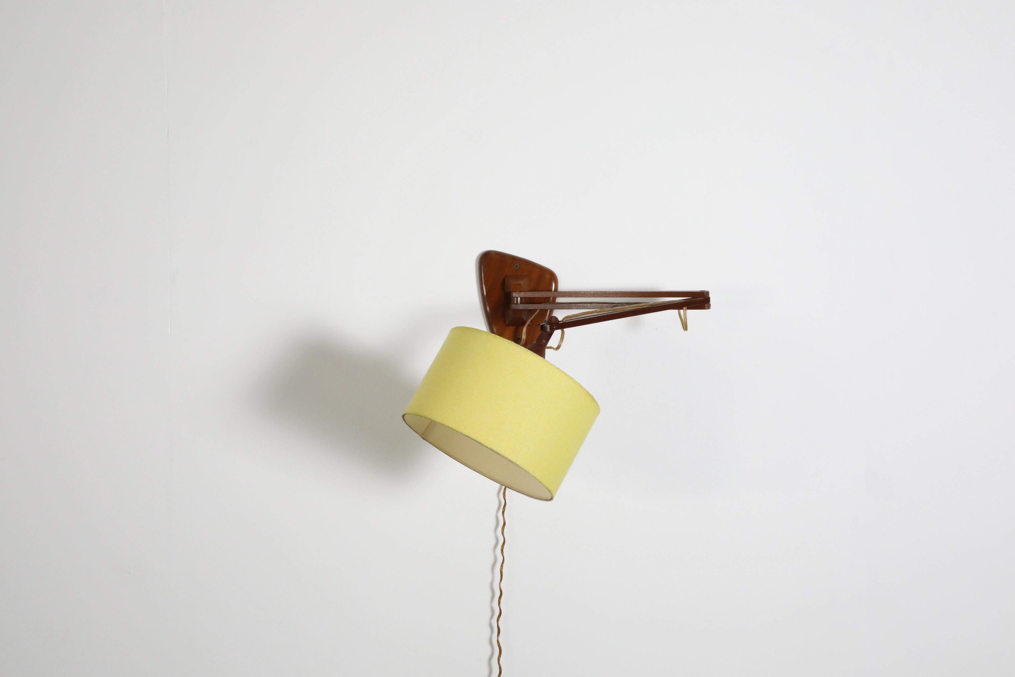 Aplique de madera de teca de mediados de siglo, con montura triangular a juego y brazo articulado. Una lámpara regulable y funcional con pantalla de tela amarilla e interruptor de botón blanco. En estado original con desgaste visible acorde con su