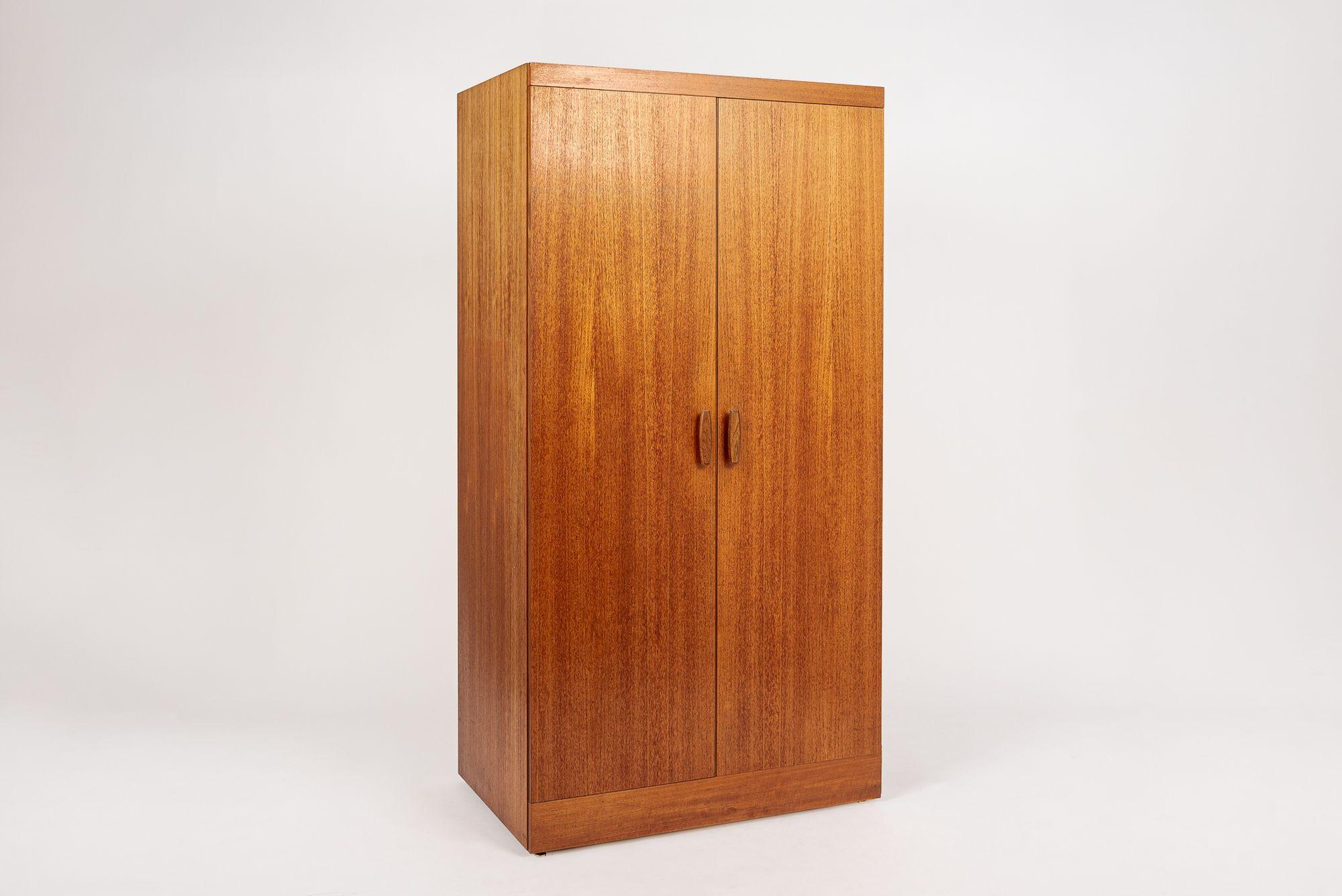 This vintage mid century modern teak wood wardrobe or armoire closet cabinet was made by G-Plan in Great Britain circa 1960. The center double doors open on spacious storage with one hanging rod across the top. This versatile, free-standing wardrobe