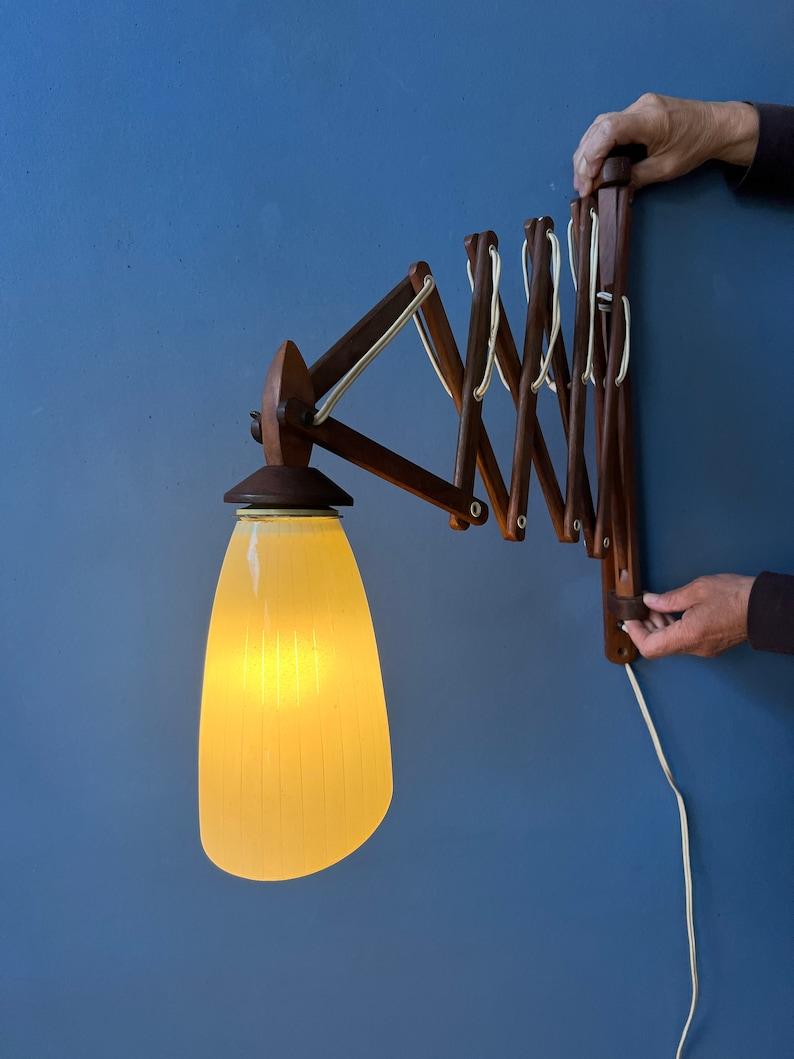 Danish mid century teak wood wall lamp with glass shade. The scissor arm mechanism is not only visually intriguing with its minimalist design but also highly functional. Effortlessly adjust the arm's position to direct light where you need it most,