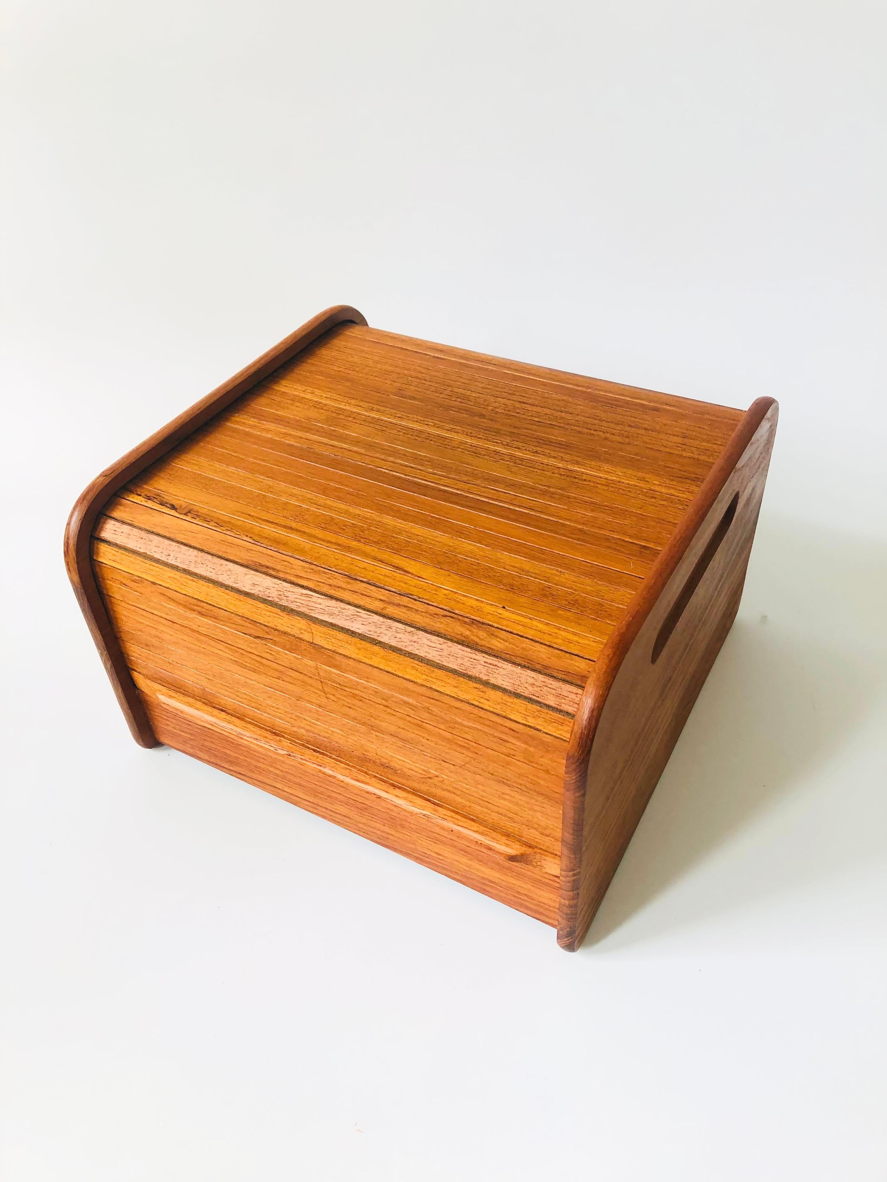 A mid century teak tambour door storage box. Features a pocketing lid that rolls back to reveal a roomy storage compartment. A single wood divider runs through the center. A stylish box for keeping odd and ends out of sight.