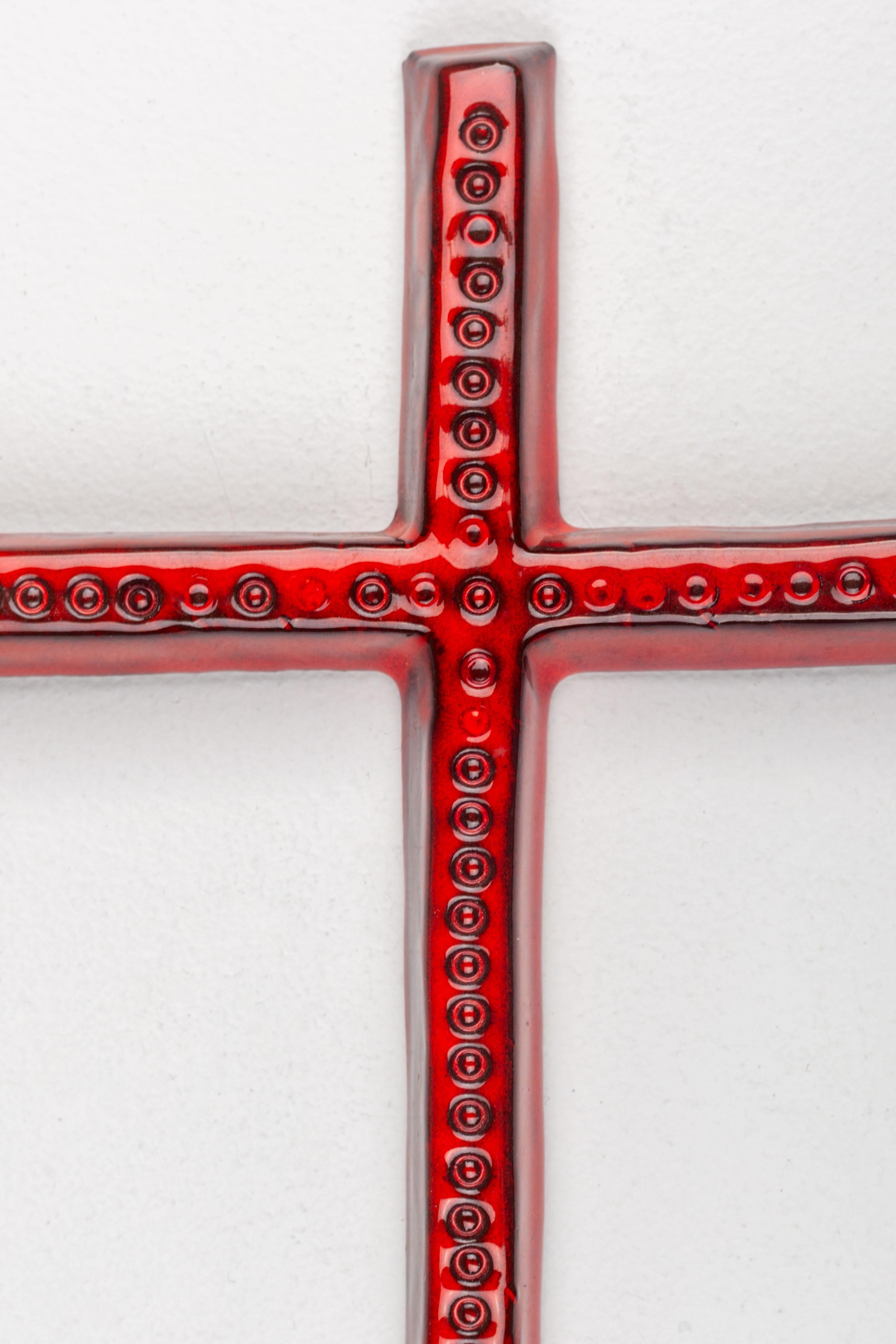 Cette croix murale moderniste en céramique European Studio Pottery se caractérise par sa forme élancée et sa finition rouge brillante, ornée d'un motif de cercles en relief qui s'étend sur toute la face de la croix, rappelant des éléments du