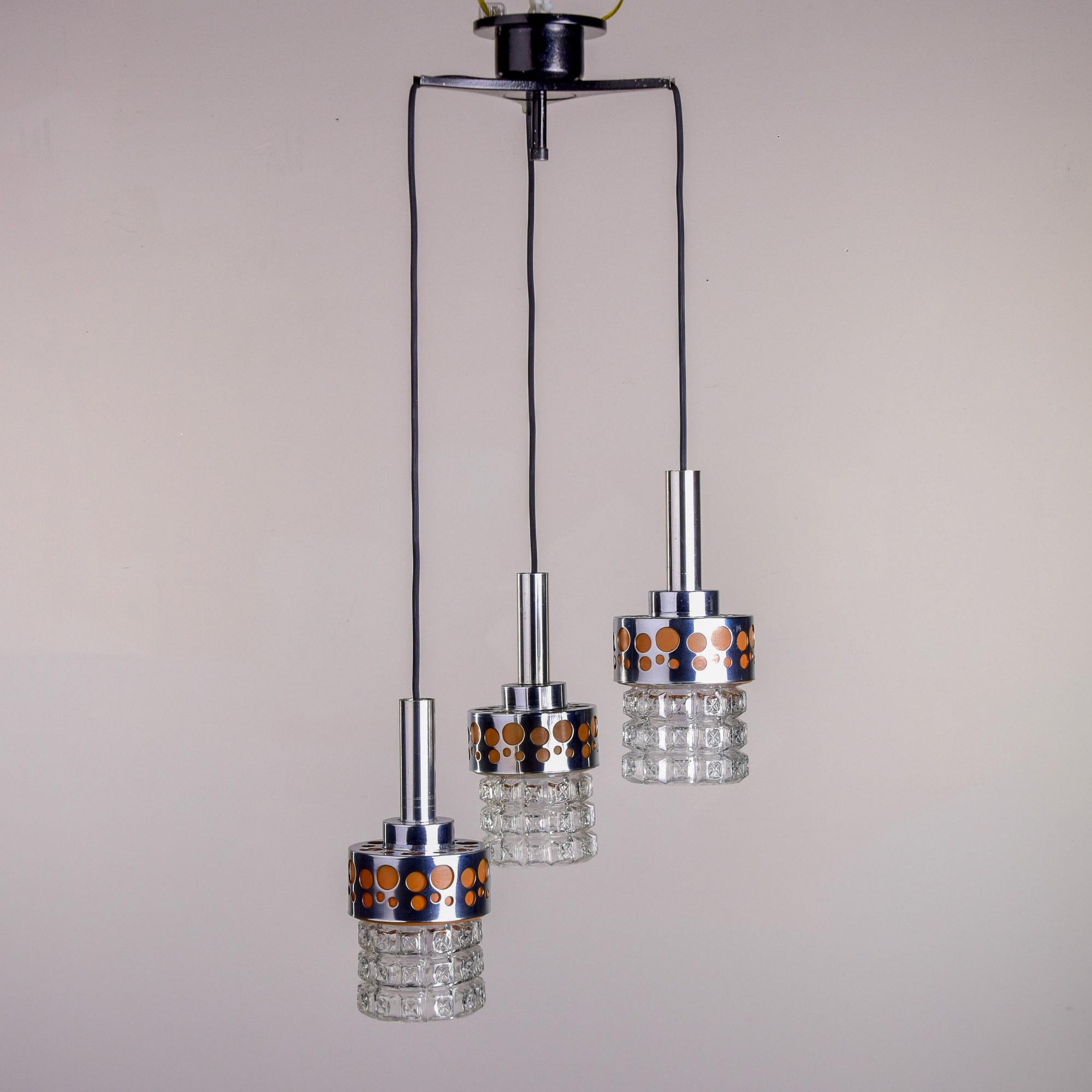 Questo apparecchio a tre luci, trovato in Italia, risale agli anni Sessanta. I tre paralumi in vetro modellato sono dotati di accessori in metallo lucido e di alcuni dettagli tagliati con dettagli color albicocca lungo la fascia superiore dei