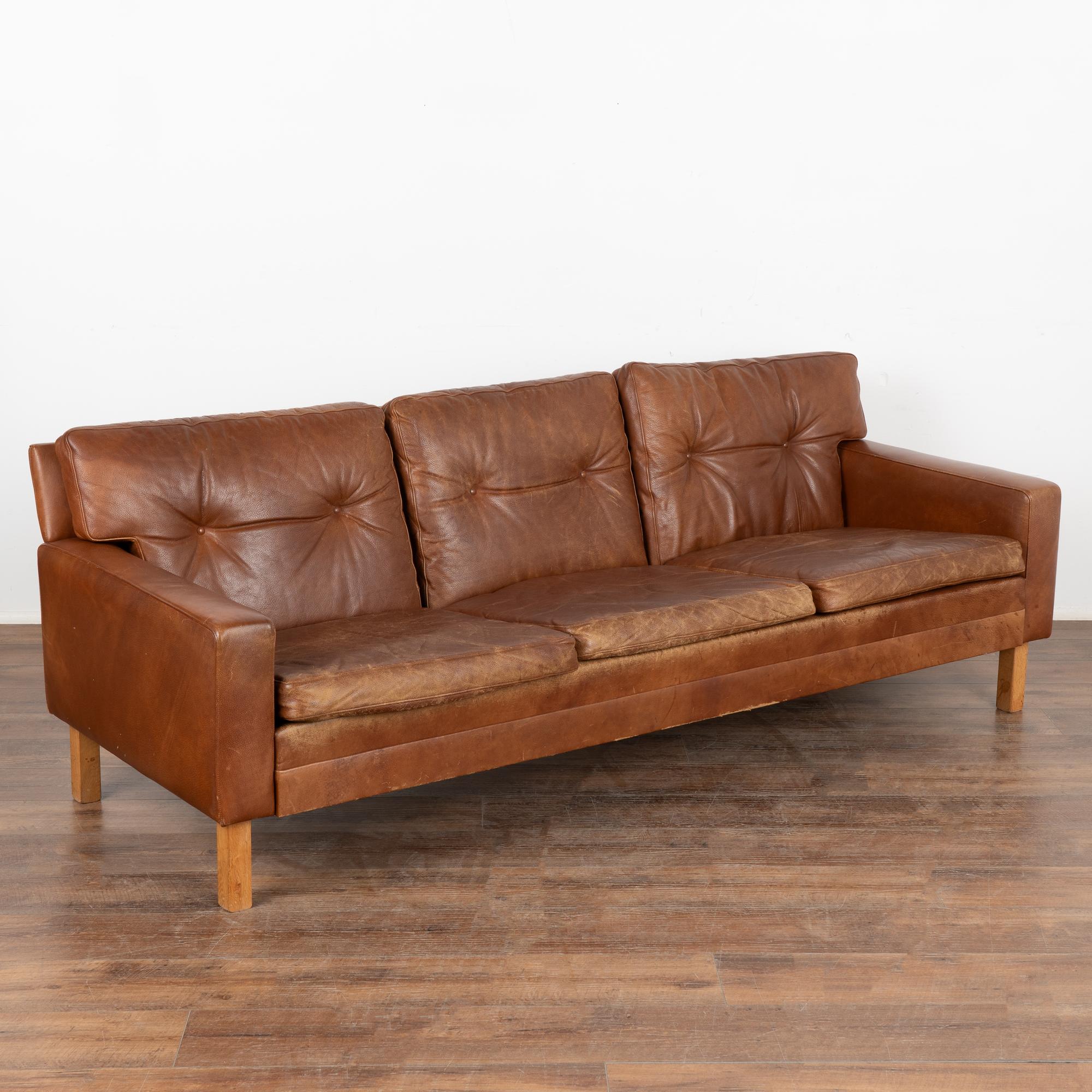 Mid-century modern brown leather three-seat sofa with button tufted back and loose seat cushions. No makers mark.
The years of use are revealed in the aged patina of the leather, including impressions, scuffs/scratches, discolorations (especially