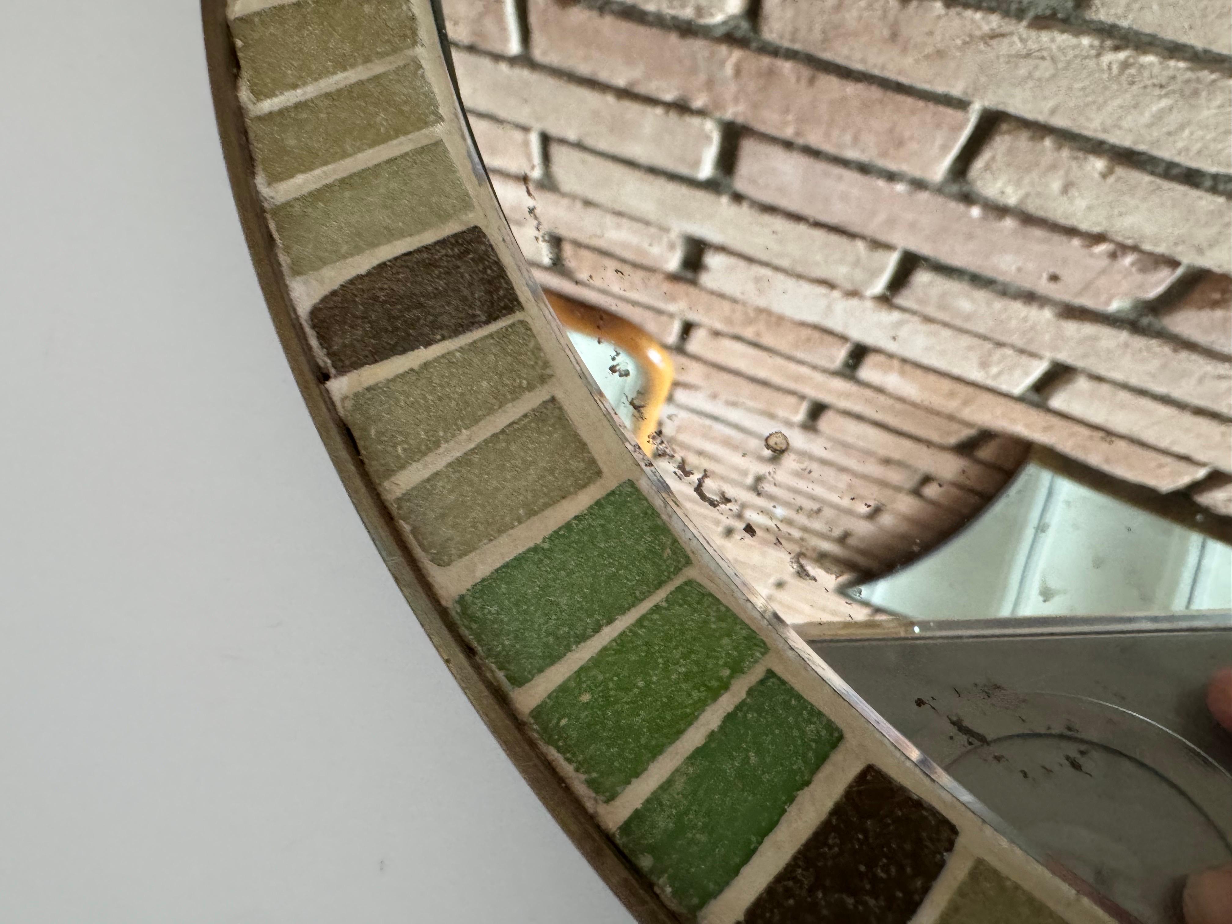 Elegante e sobrio specchio da parete rotondo con intarsio di piastrelle di vetro e una splendida cornice in ottone invecchiato. Lo specchio presenta lievi macchie di perdita di argento intorno al bordo e i colori delle piastrelle includono due