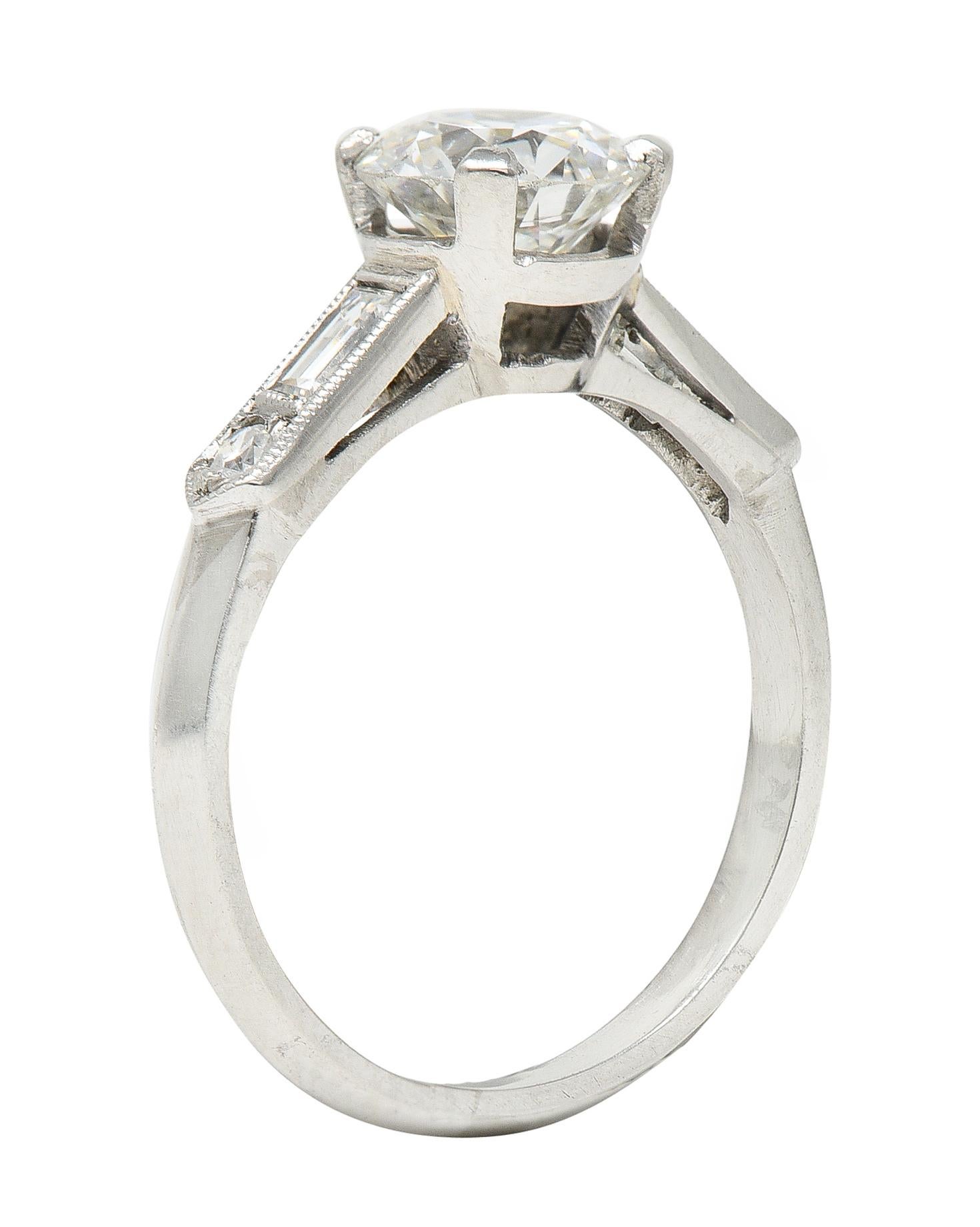 Engagement ring centers a round transitional cut diamond weighing 1.25 carat total - H color with VS1 clarity. Prong set in basket - flanked by cathedral shoulders with flush set baguette and single cut diamonds. Weighing approximately 0.35 carat