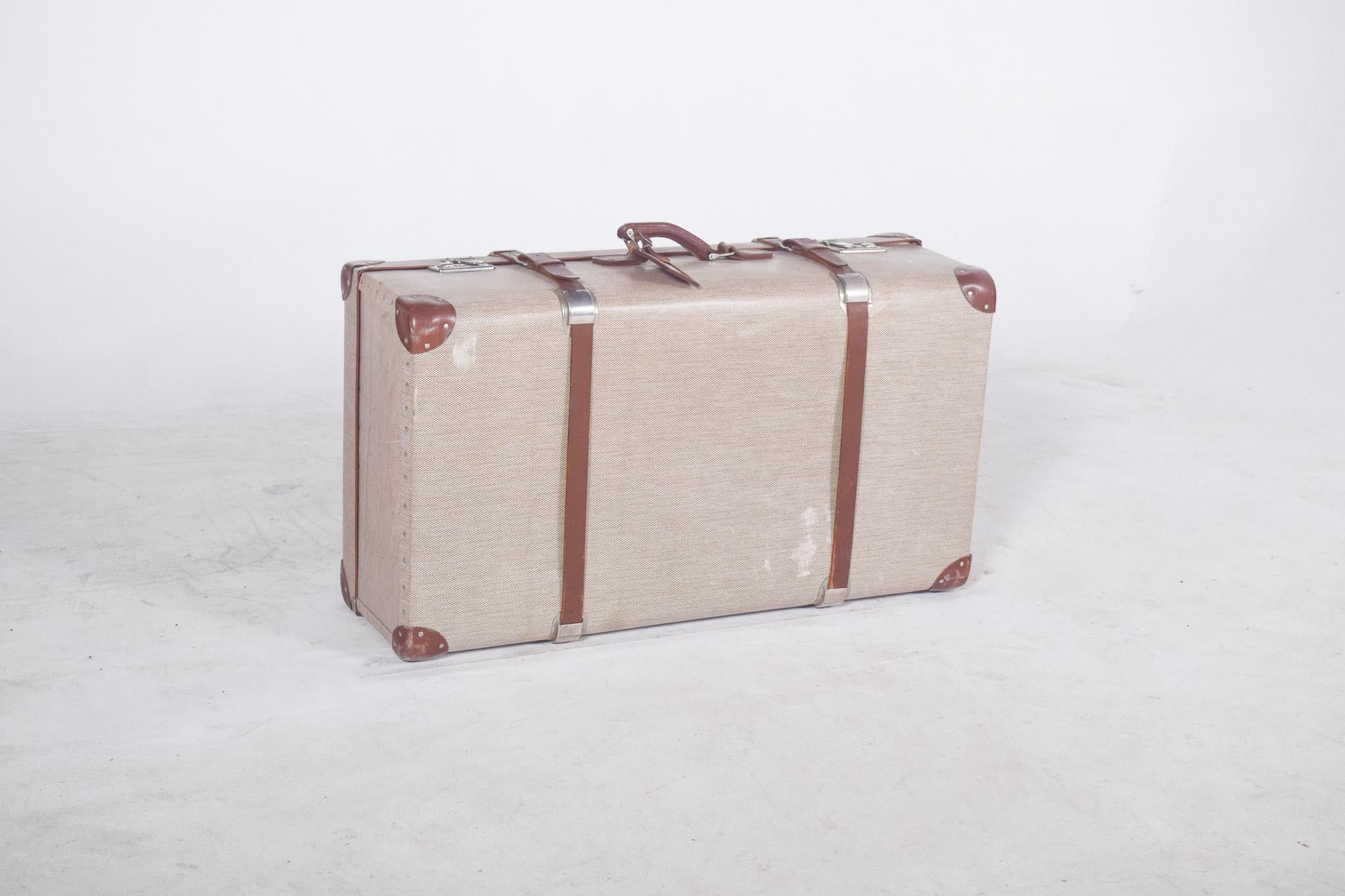 Sac de voyage en cuir. De forme rectangulaire, avec une structure rigide et des proportions généreuses, il présente une doublure en tissu acrylique beige à motifs discrets, avec des traces d'utilisation visibles.
Les coins sont renforcés par des