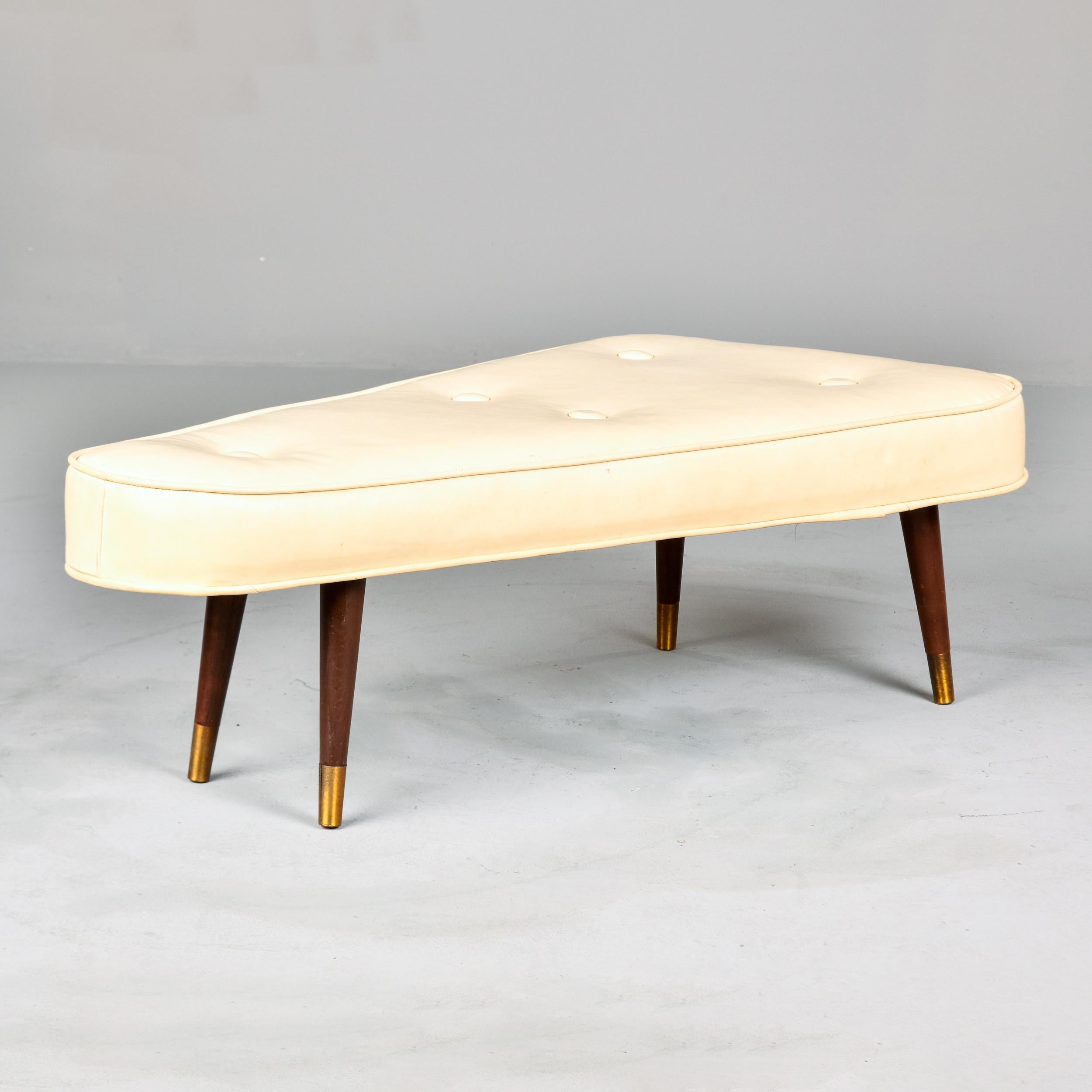 Found in the US, this triangular bench or stool dates from the late 1950s or early 1960s. Elongated triangular bench seat is upholstered in cream-colored vinyl with button tufting, single welt trim and four brass-tipped tapered wood legs. Unknown