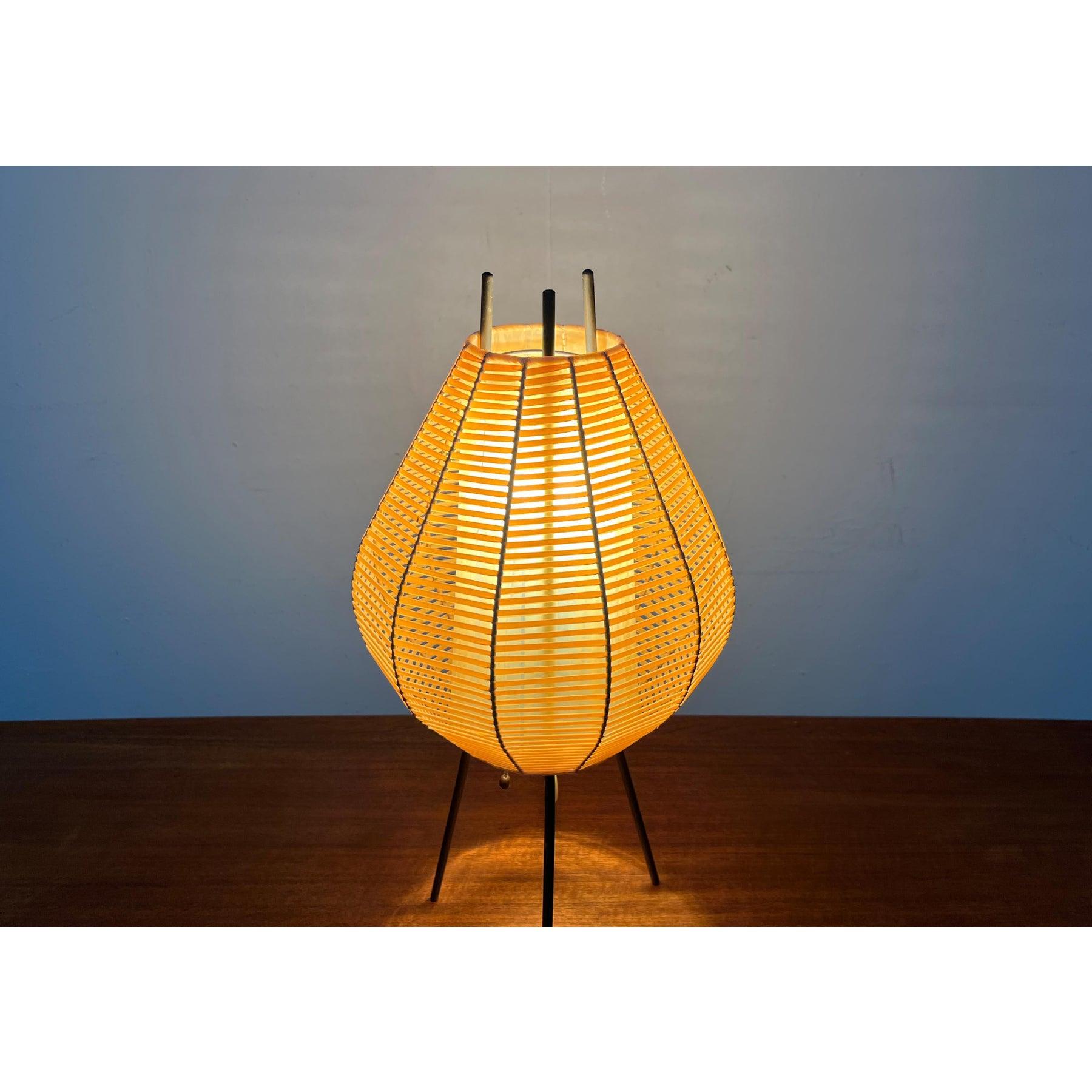 Seltene Tischlampe aus der Mitte des Jahrhunderts mit einer charakteristischen Schirmform.

Der markante Lampenschirm wird von einem inneren Gestell getragen, das unten in ein Dreibein übergeht. Die Stäbe des Stützrahmens schauen oben heraus. Der