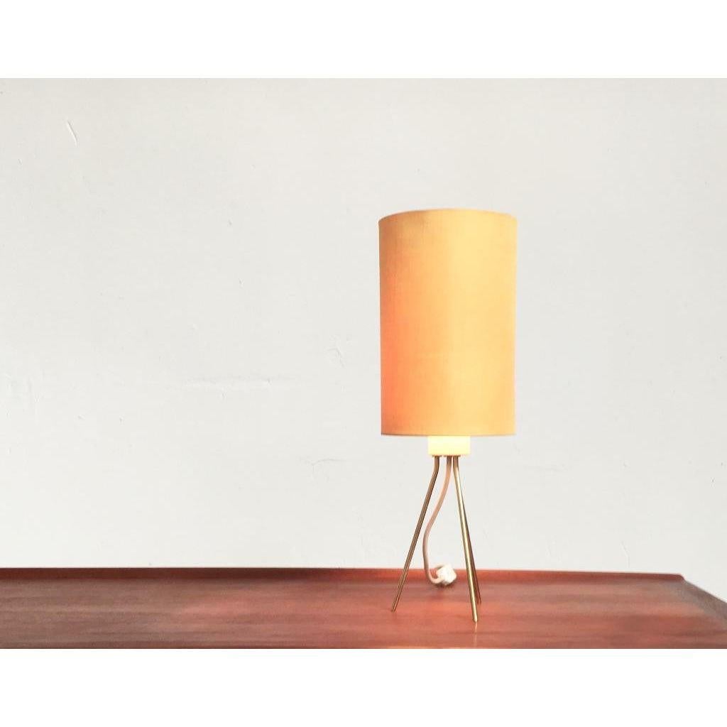 Mid Century tripod table lamp with shade.

Very reserved in terms of shape and color. The lamp balances on 3 brass-colored feet. The shade is cylindrical and covered on the outside with a yellow textile. The light has a cord switch.
The condition is