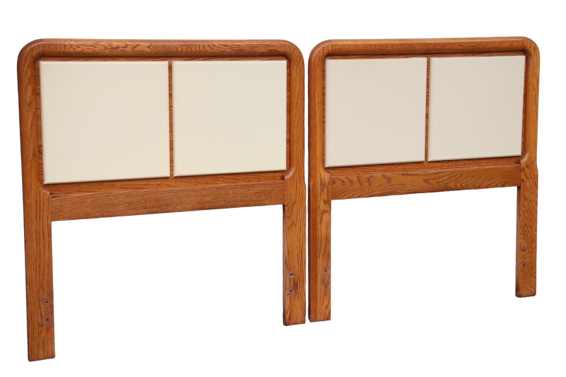 A pair of twin size mid-century headboards made of oak. Each has a gently curved frame, inset with two square panels painted a light cream. Dimensions per headboard.
