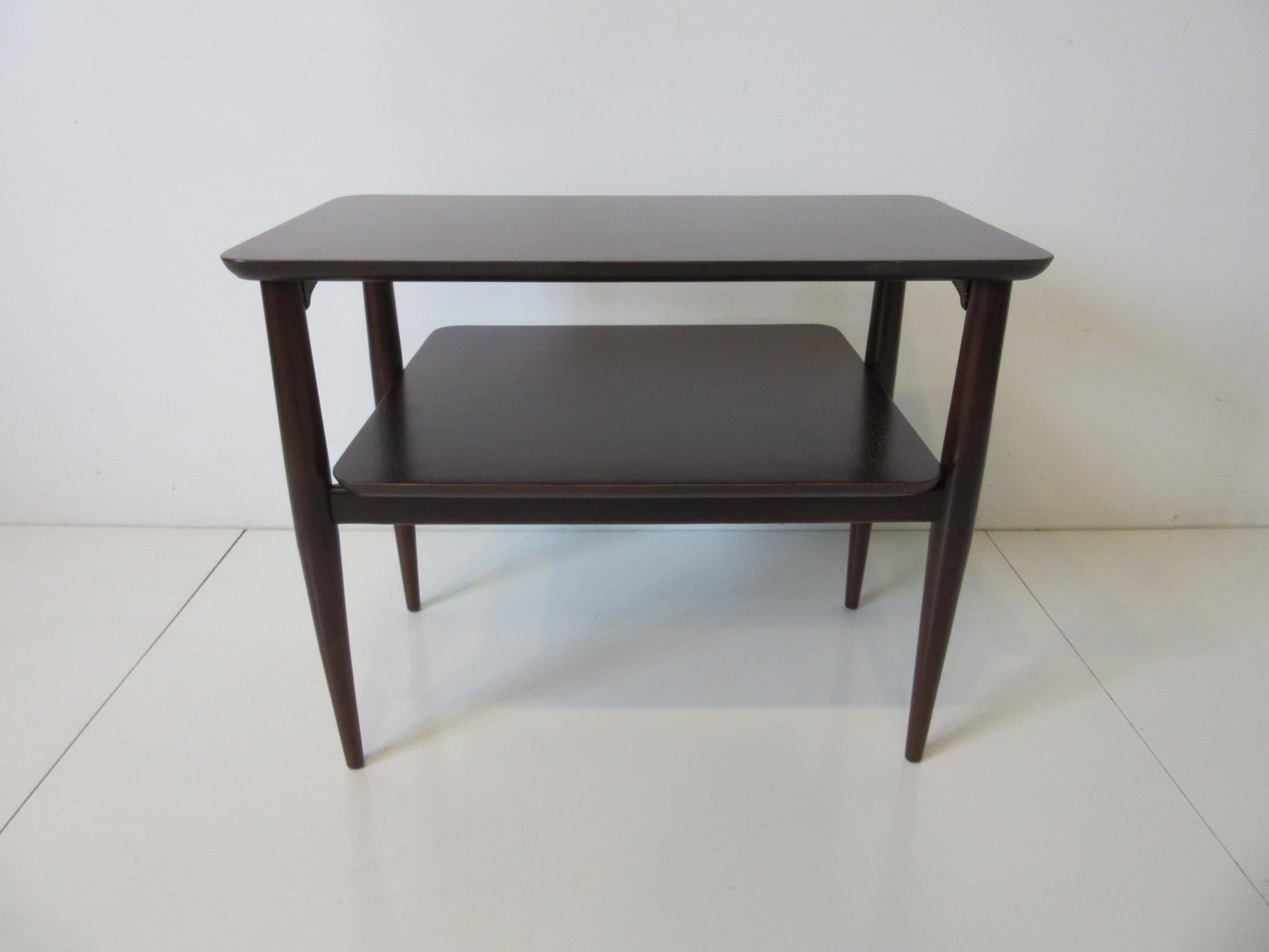 A nice sized midcentury two-tiered wood side table with beveled edges and conical legs refinished in a rich ebony tone giving the piece a very high stylish feel. The perfect table to share between two lounge chairs or able to stand alone.
 
